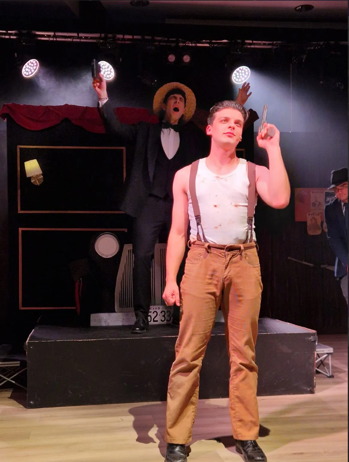 Stage scene from a theatrical performance featuring two male actors. One actor in the foreground wears a white tank top, suspenders, and tan pants, holding a gun in his right hand. The other actor in the background, dressed in a suit with a straw hat