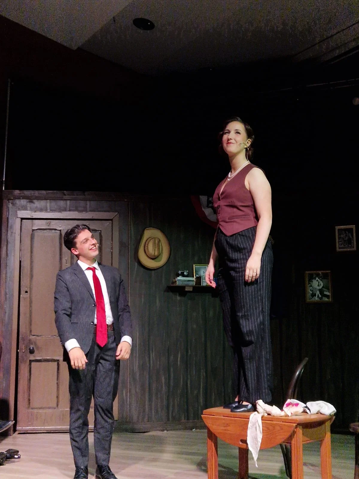 A woman standing on a table and a man in a suit with a red tie looking at her, inside a vintage-style room with wooden walls and framed photos.
