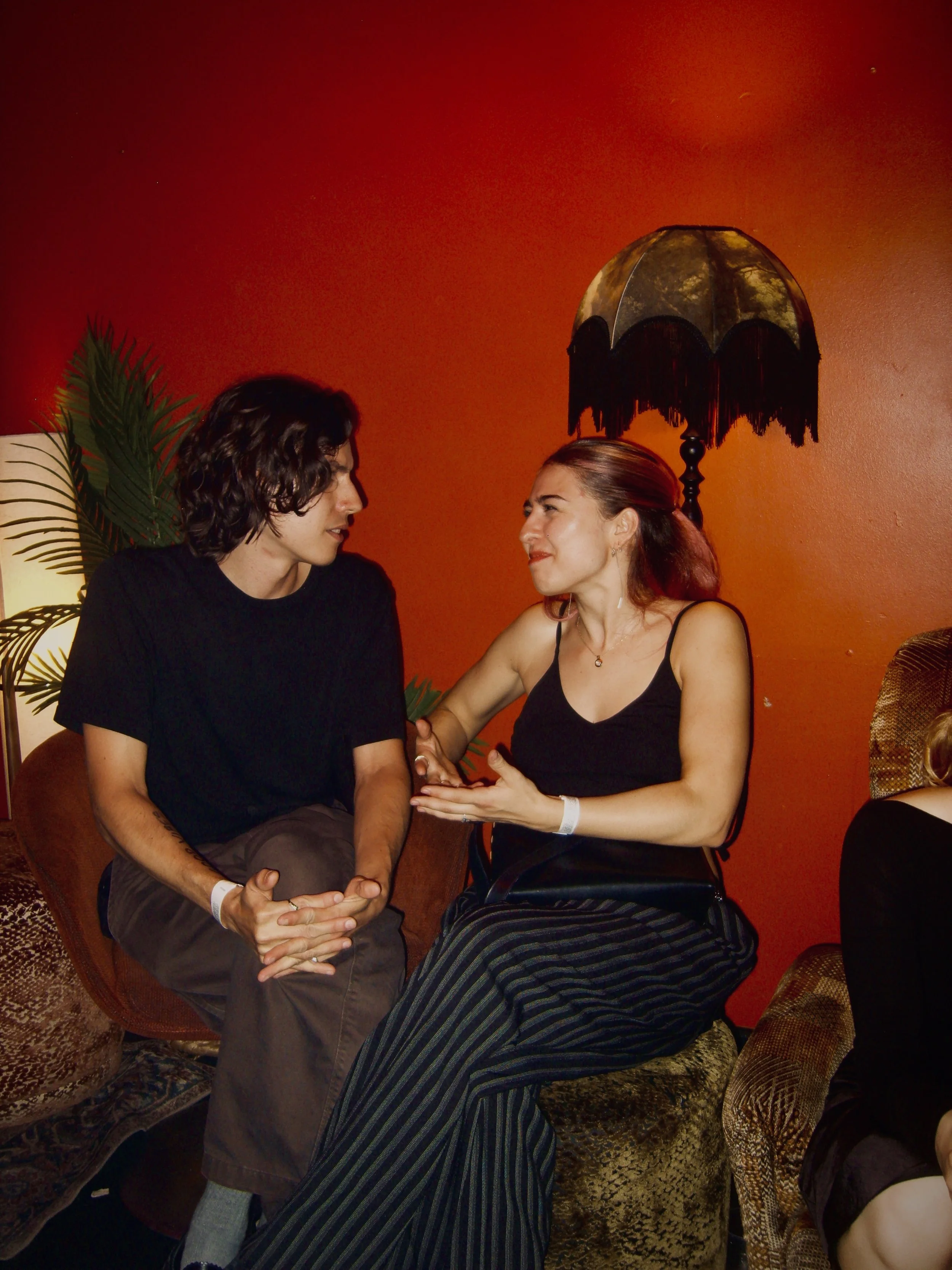 A man and woman engage in an intimate conversation while holding hands, sitting close together in a cozy room with red walls, decorated with a black lamp with a tassel fringe and a leafy plant.