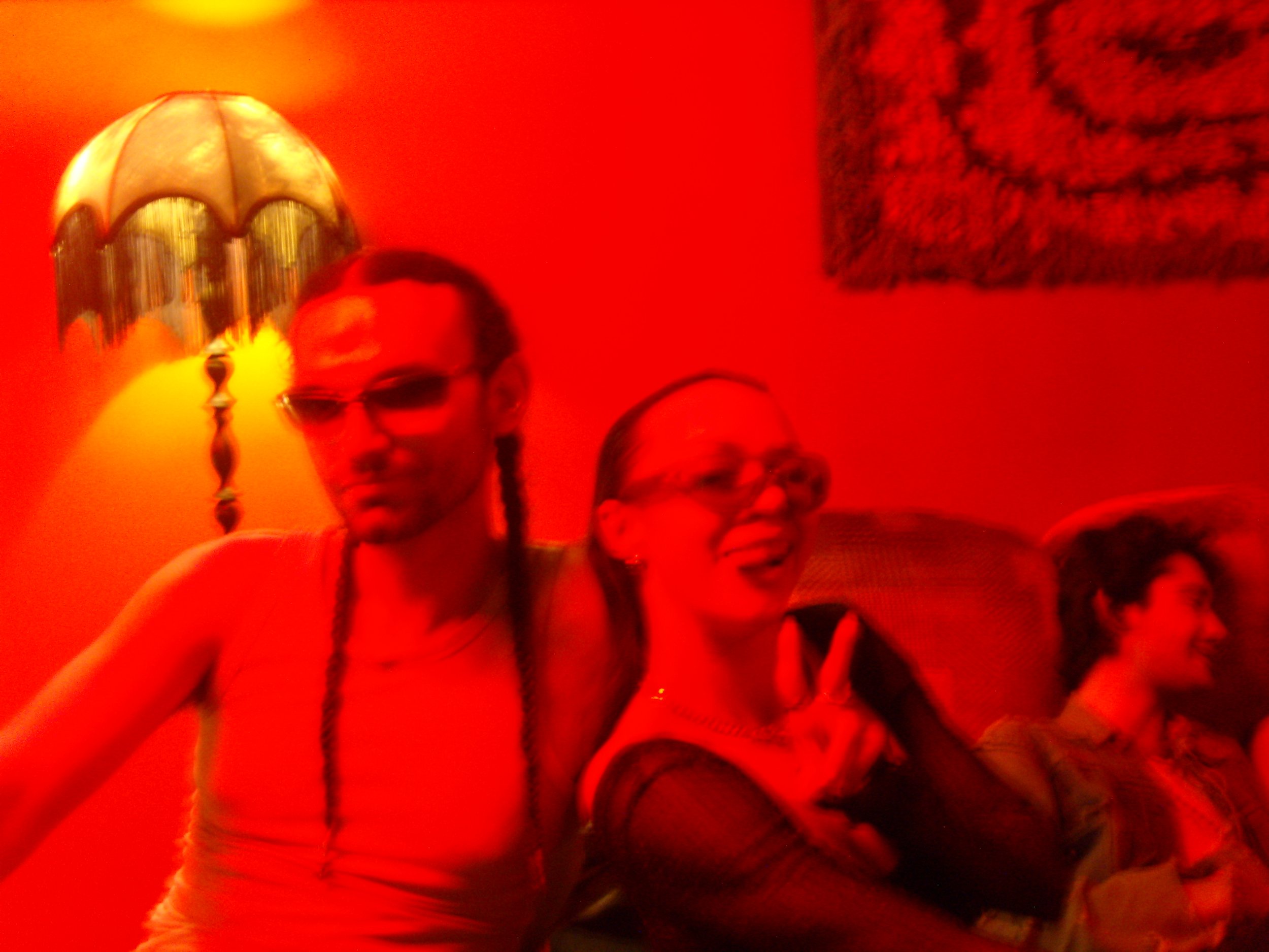 Group of people at a party with red lighting, one person in the foreground wearing glasses and a beaded necklace.