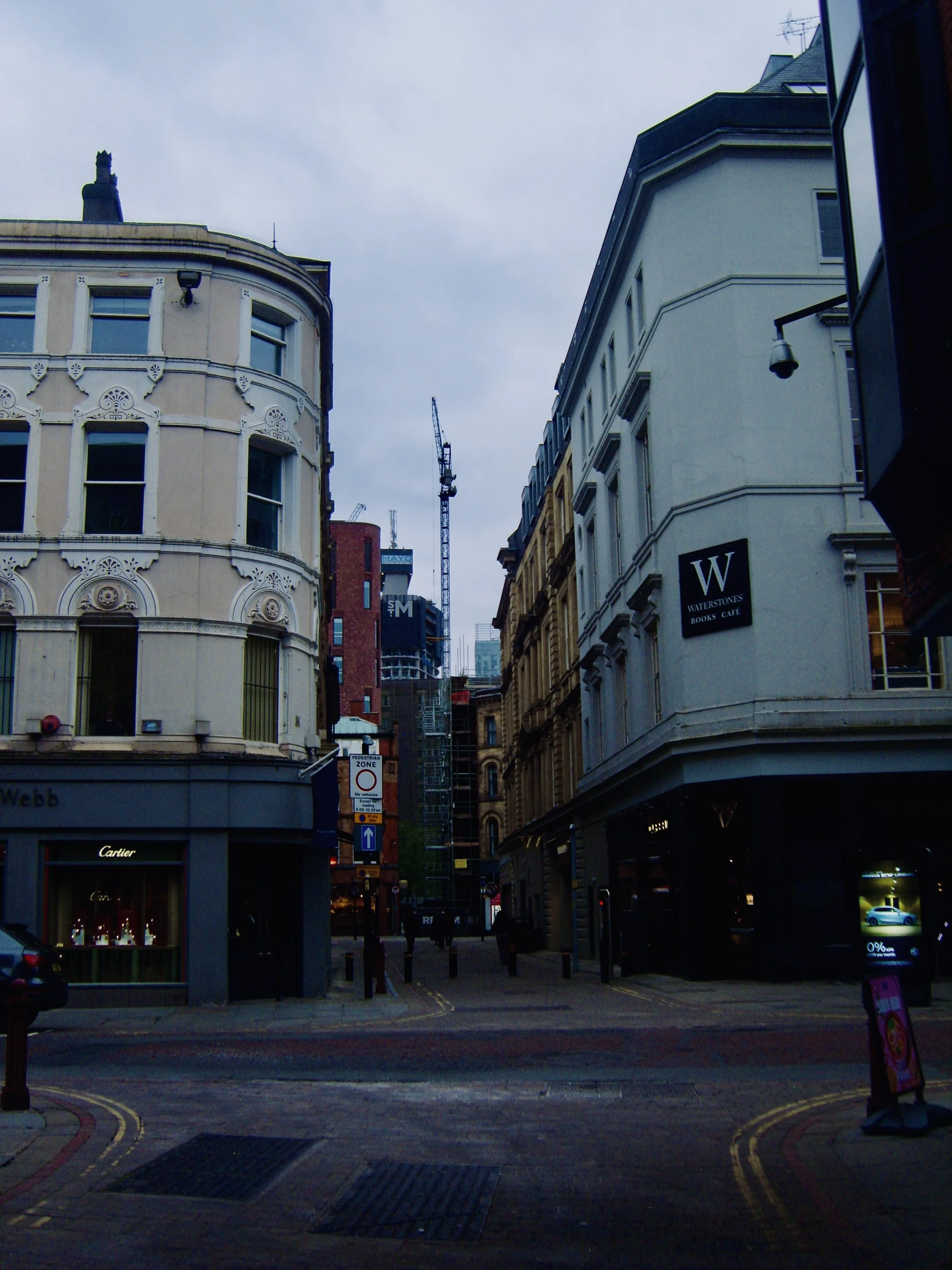 A city street corner with shops, including Cartier, and a pedestrian zone sign. Buildings on both sides and cranes in the background under a cloudy sky.
