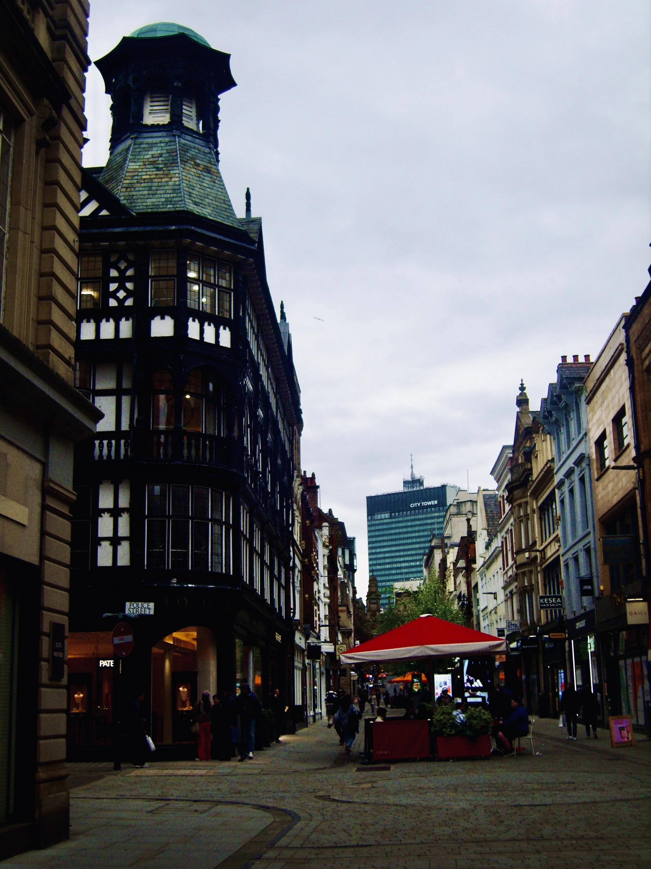 A street scene with historic and modern buildings, people walking, outdoor cafes, and a cloudy sky.