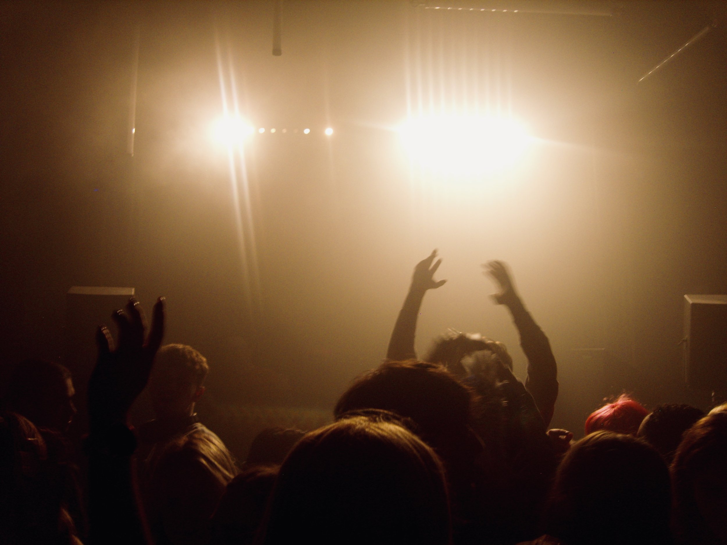 A crowd of people at a concert with raised hands, bright stage lights shining in the background, and smoke or fog creating a hazy atmosphere.