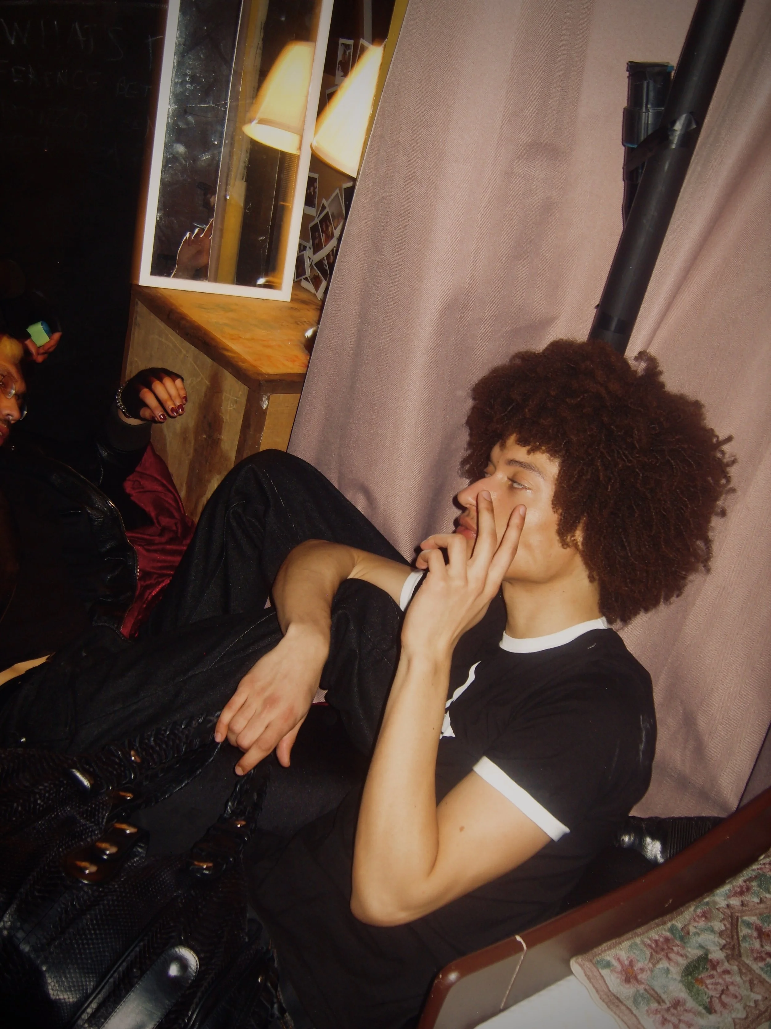 A young woman with curly hair and a black and white T-shirt listening to music with headphones while sitting on a beige couch in a room with pink walls, a wooden side table, and a black object resembling a tube or pole.