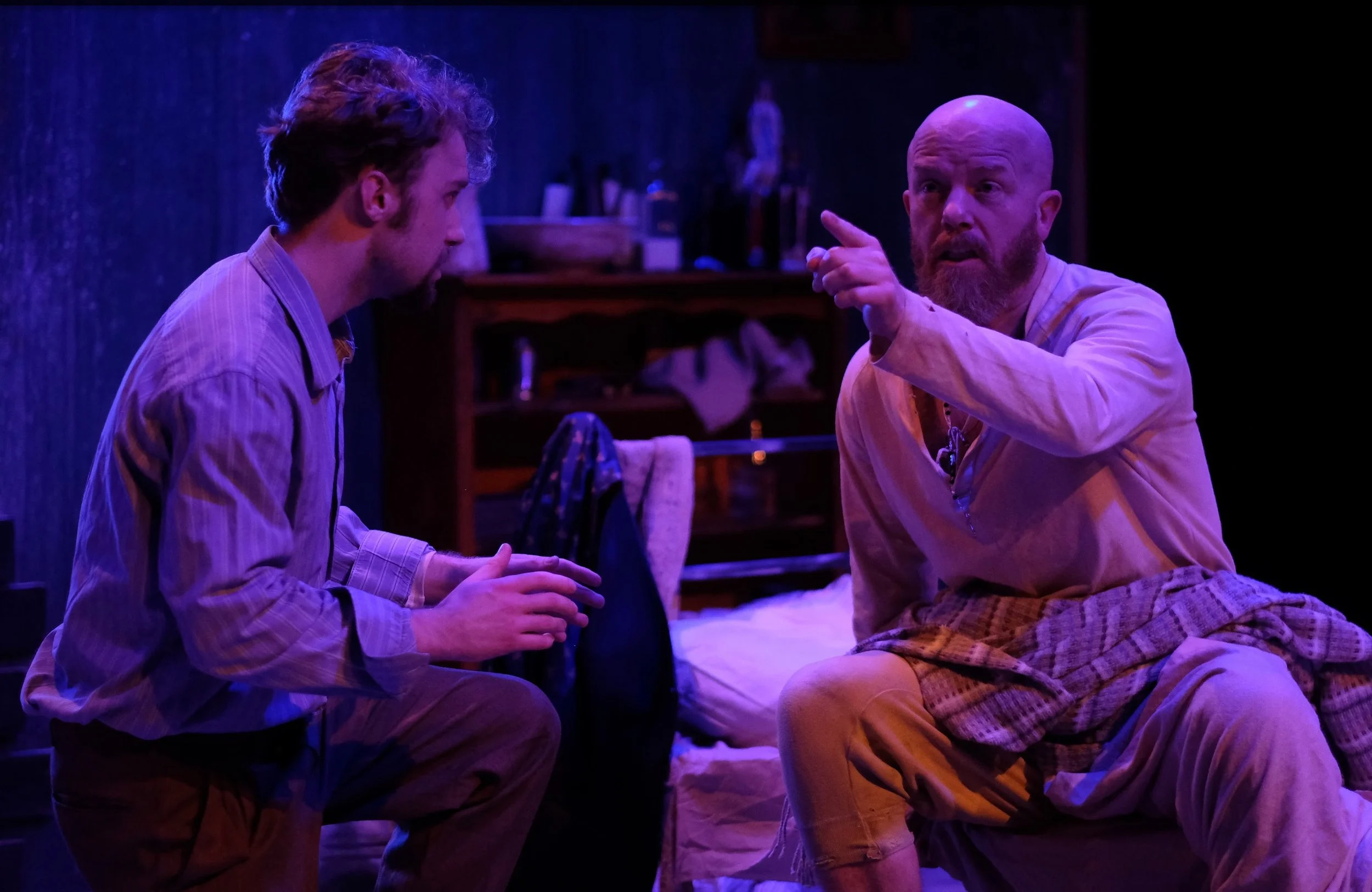 Two men having a serious conversation in a dimly lit room with blue and purple lighting, sitting close to each other, one man is pointing his finger while the other listens.