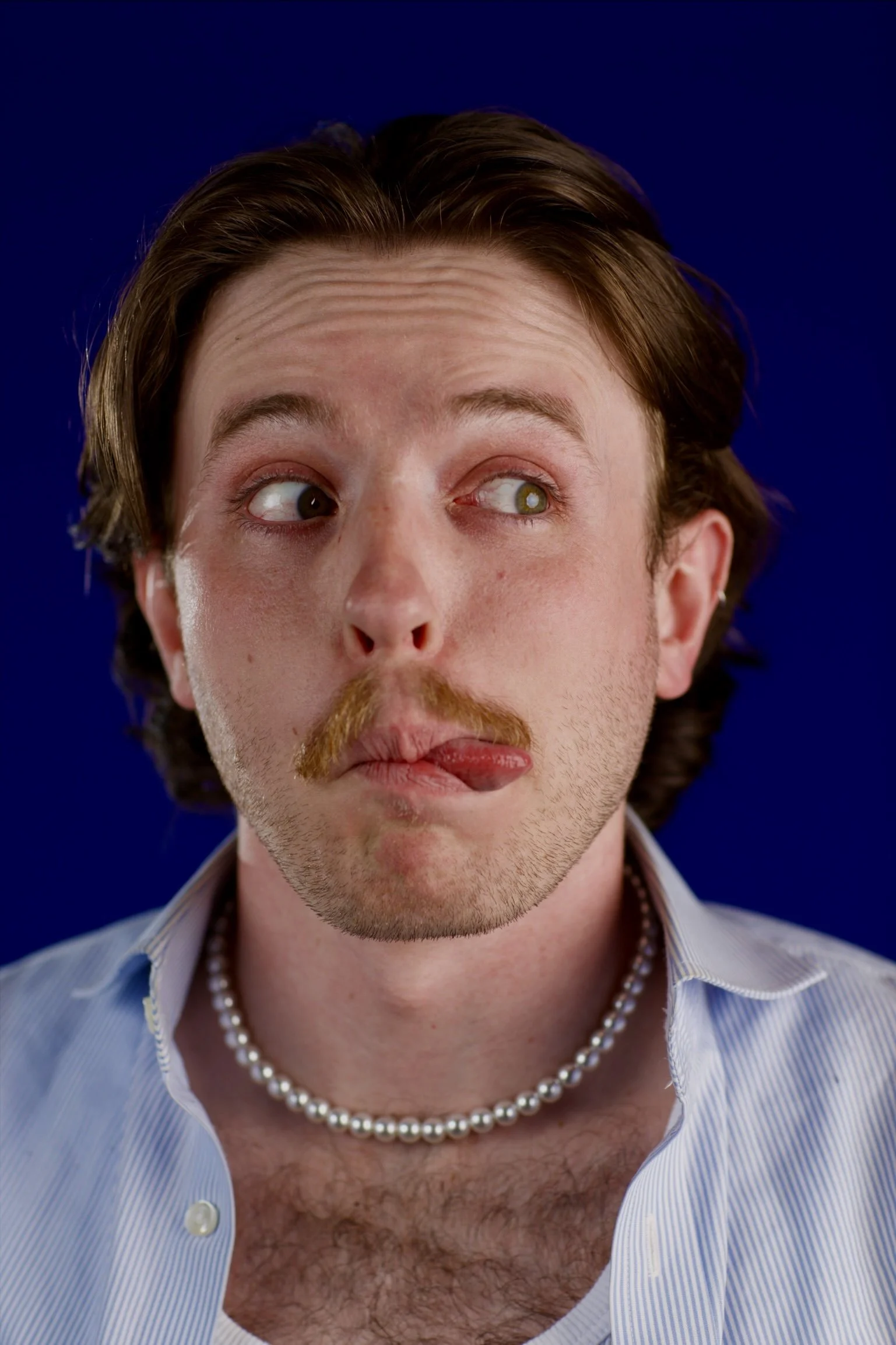Close-up of a man with brown hair, wearing a light blue striped shirt, pearl necklace, and sticking out his tongue with a playful expression.