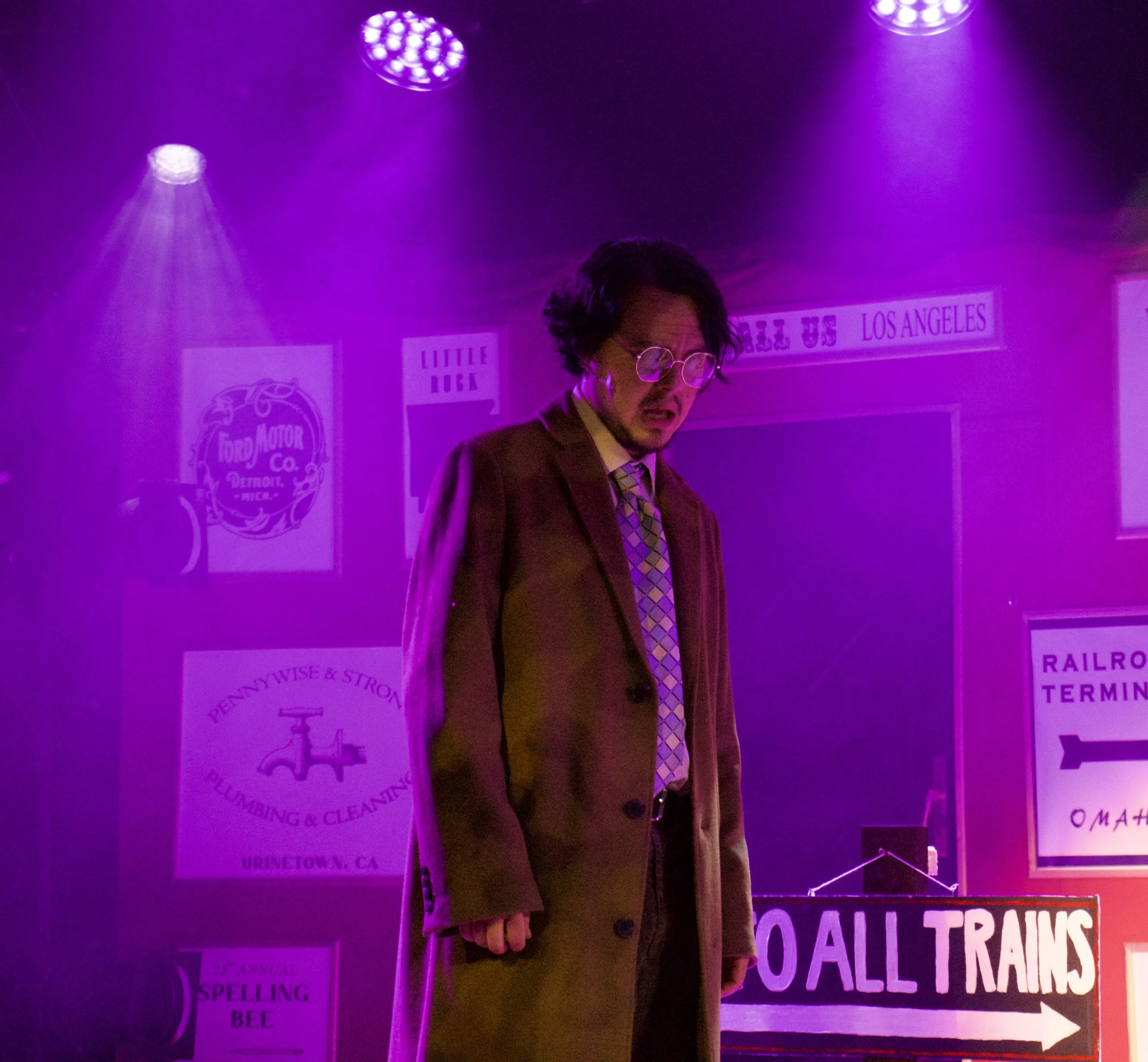 A man with dark, curly hair and glasses wears a suit and patterned tie, standing in a purple-lit room with signs and posters about Los Angeles and other city-related topics.