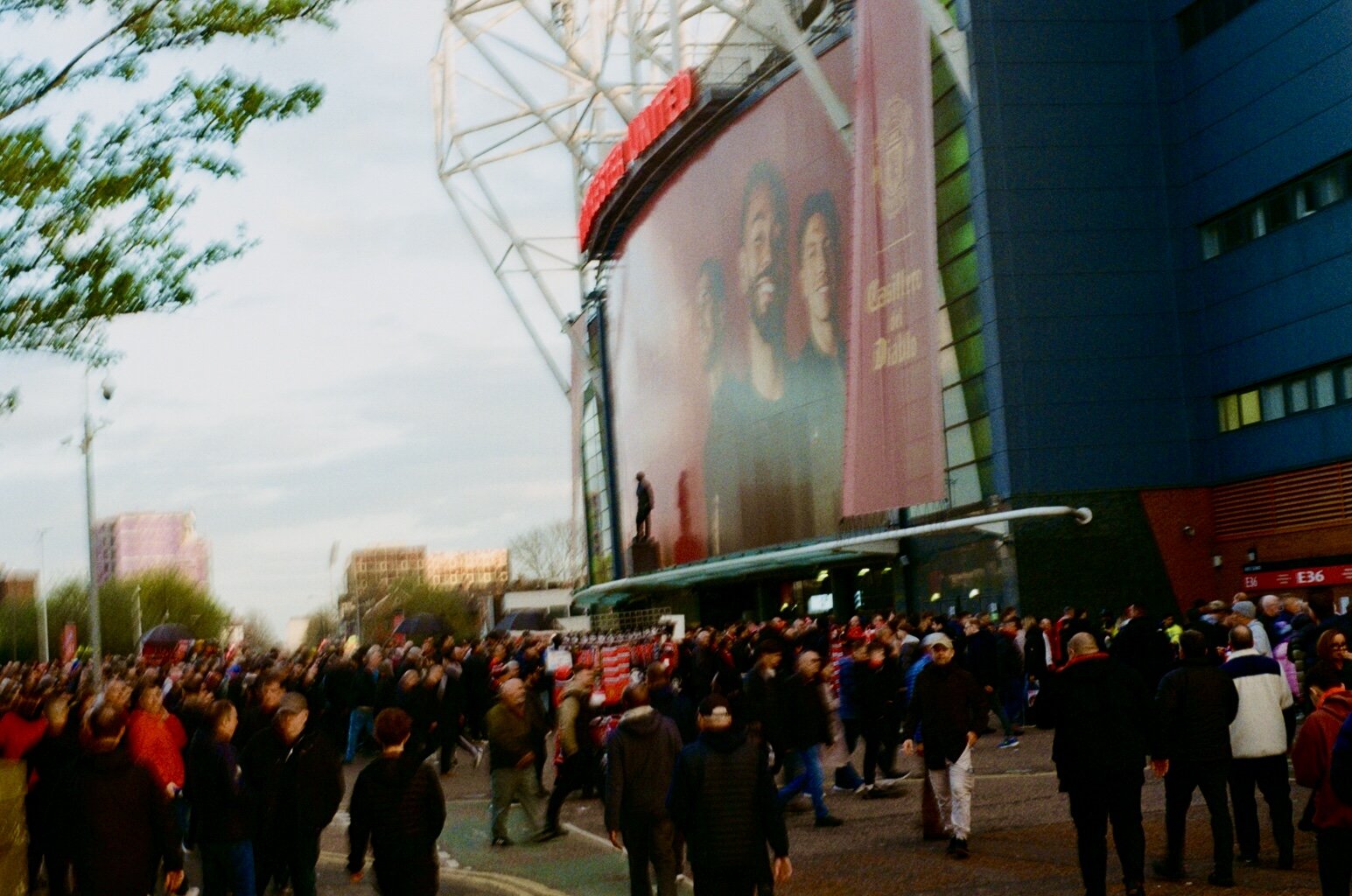 Crowd of people gathering outside a large modern building with digital advertising screens, in an urban area during daytime.