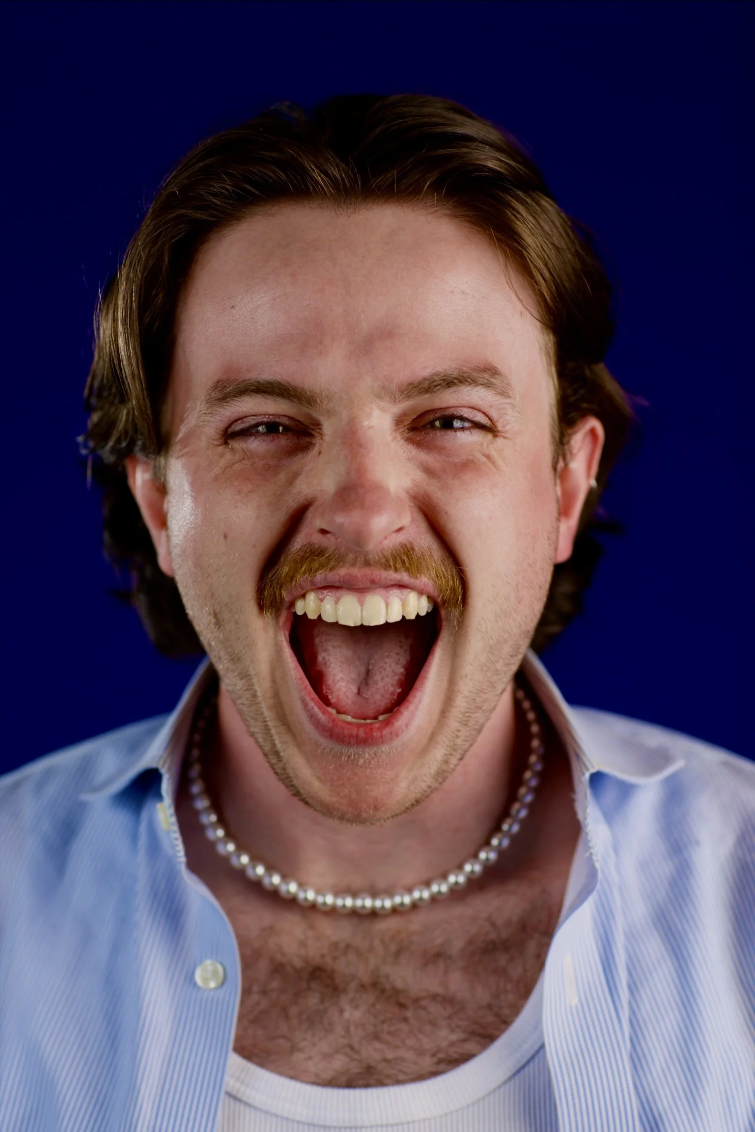 A man with brown hair, a mustache, and a beard, smiling widely with his mouth open, wearing a blue and white striped shirt, a pearl necklace, and a white shirt underneath, against a dark blue background.