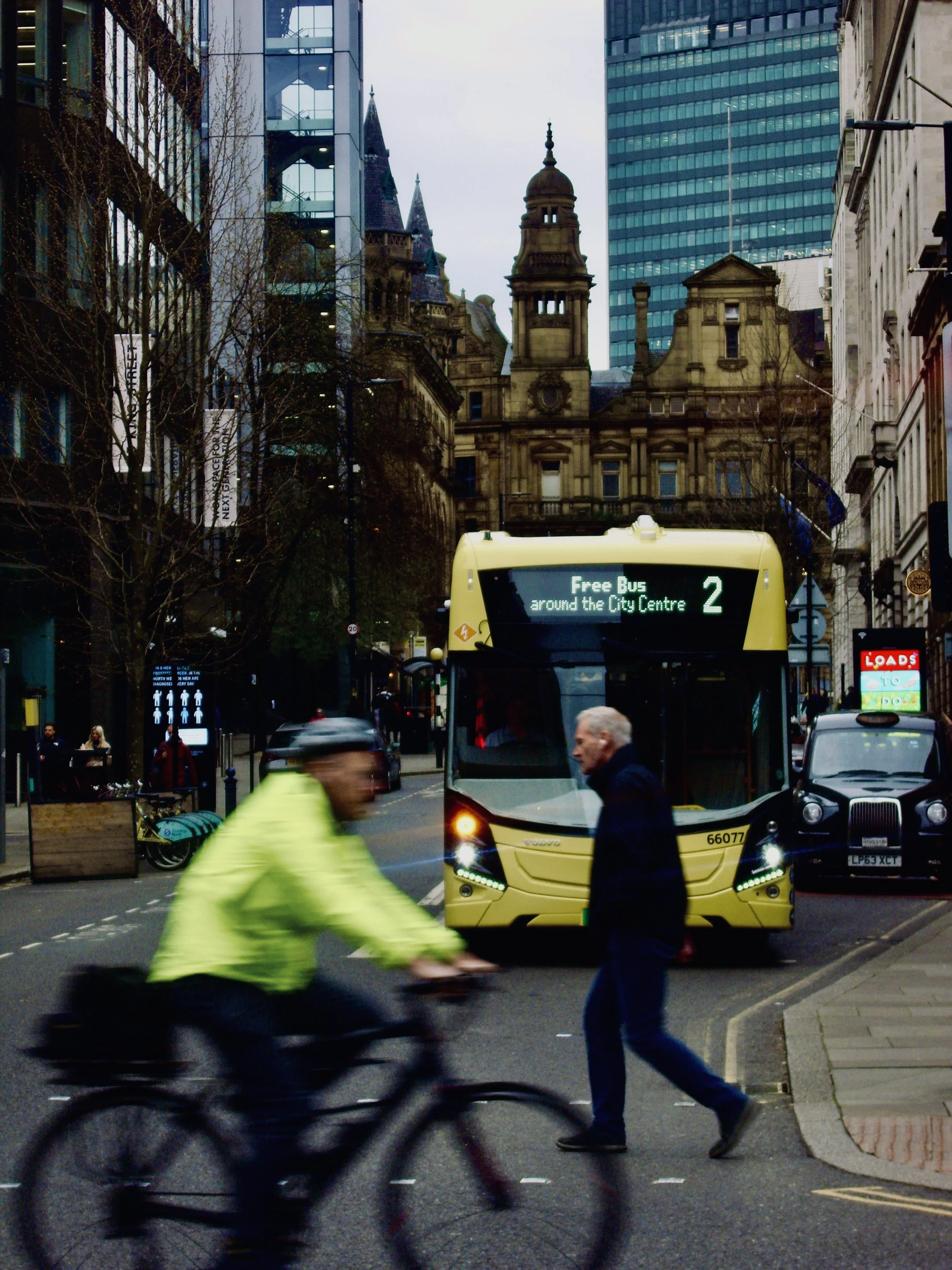 A busy city street scene with pedestrians, a bus, and a cyclist in motion. The background features modern glass buildings and historic architecture.