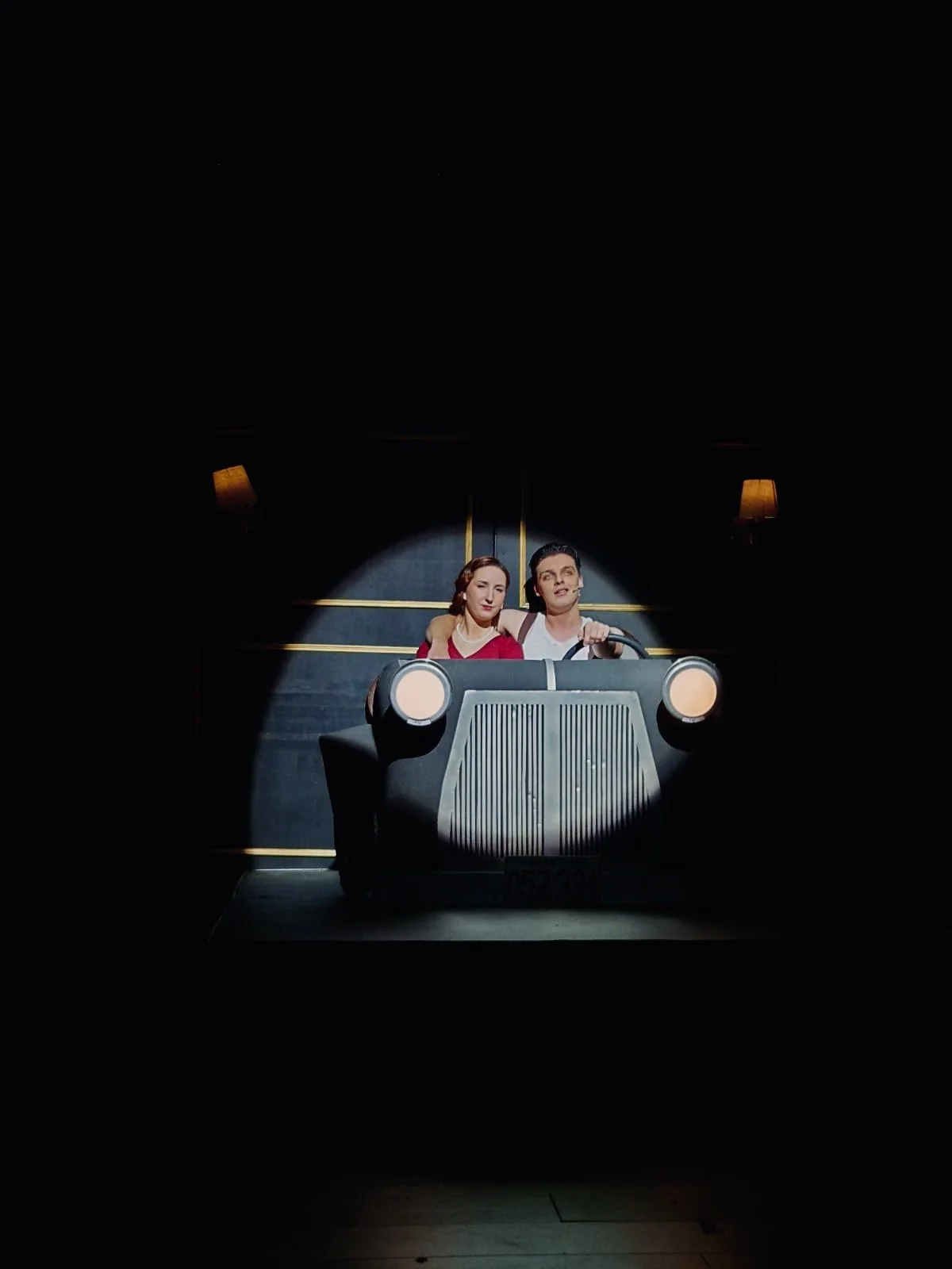 Scene from a stage play showing a man and woman sitting in a vintage car prop, spotlight on them, dark background with two wall-mounted lamps.