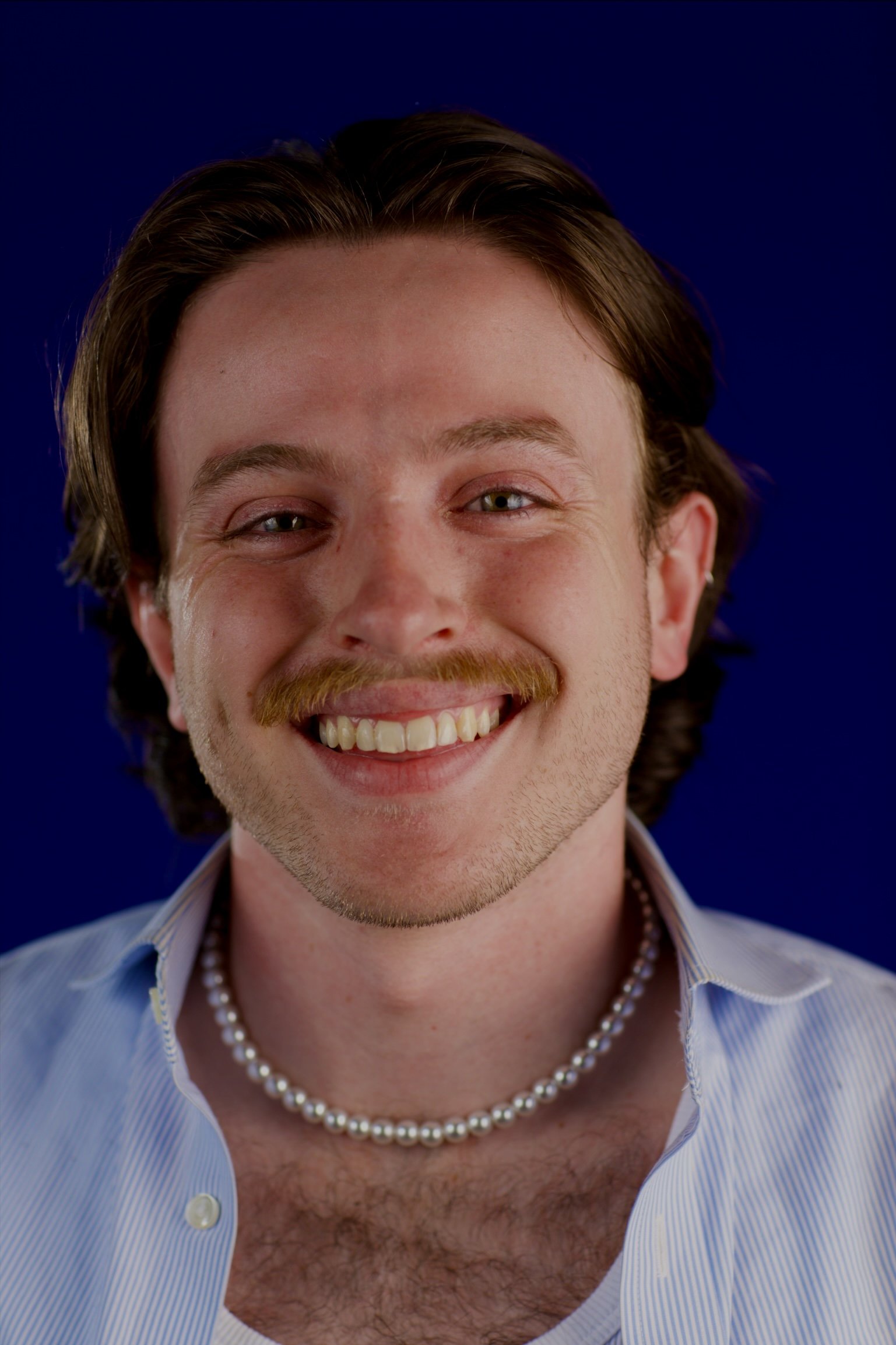 Close-up of a smiling man with light skin, brown hair, and a beard, wearing a pearl necklace, a light blue button-up shirt, and a white undershirt, against a dark blue background.
