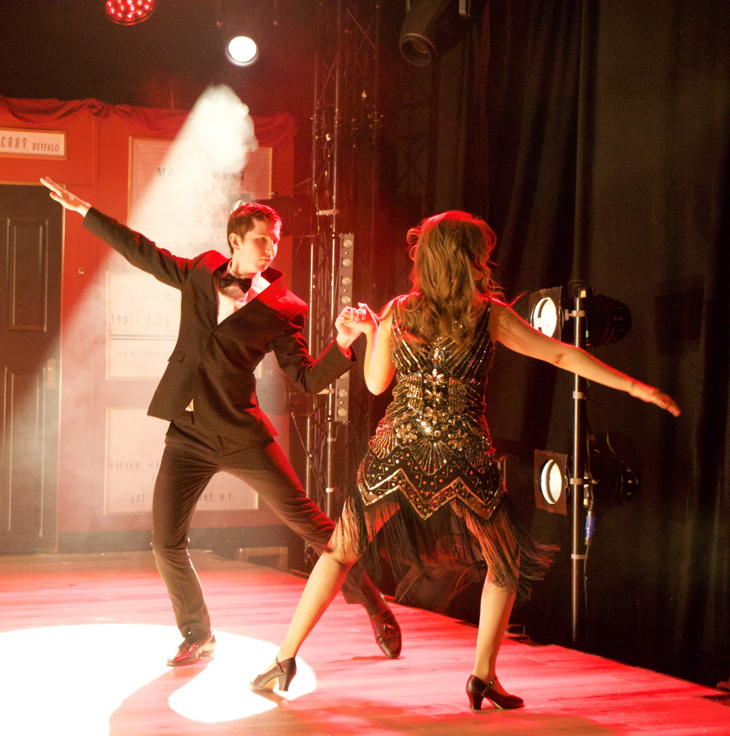 Dancers perform on stage with red and warm lighting, one man in a black tuxedo and bowtie, and a woman in a sparkling dress, captured in mid-motion
