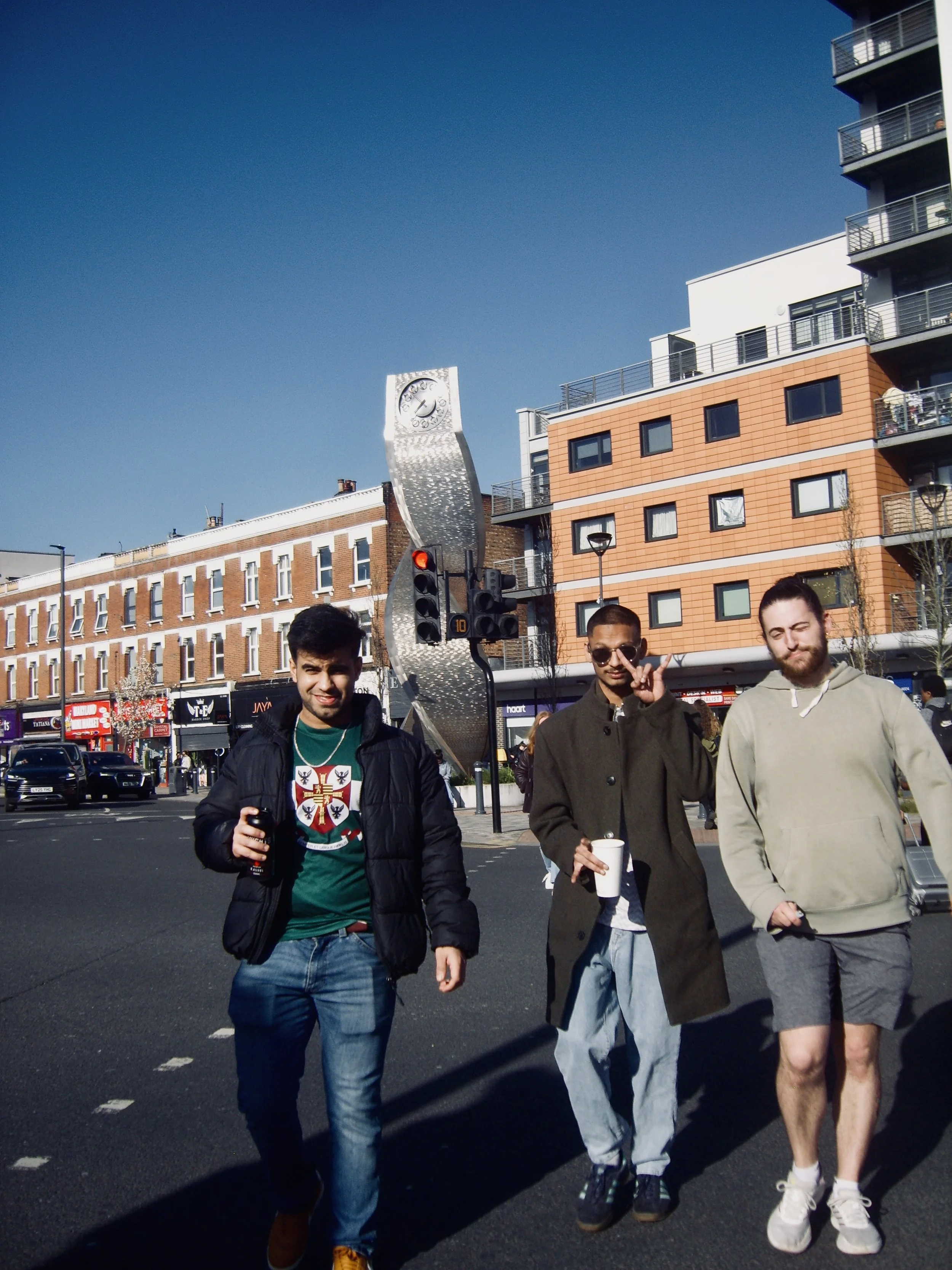 Three young men walking down a city street during daytime, with buildings, a clock sculpture, and a traffic light in the background.
