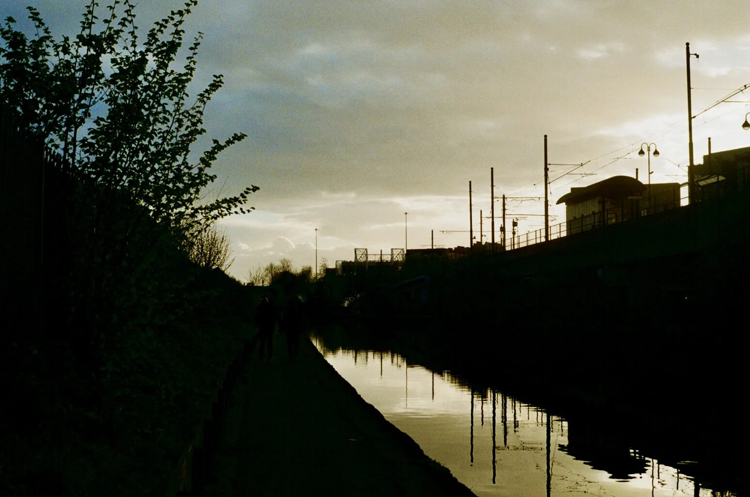 Silhouettes of two people walking along a canal with trees on the left and power lines and buildings on the right, under a cloudy sky at sunset.