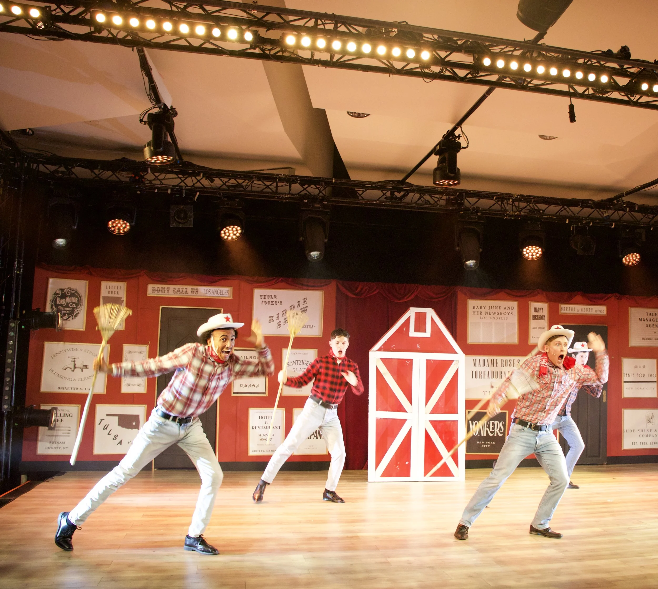 A group of performers on stage dressed in western-themed cowboy outfits, dancing and performing in front of a red barn backdrop with white trim.