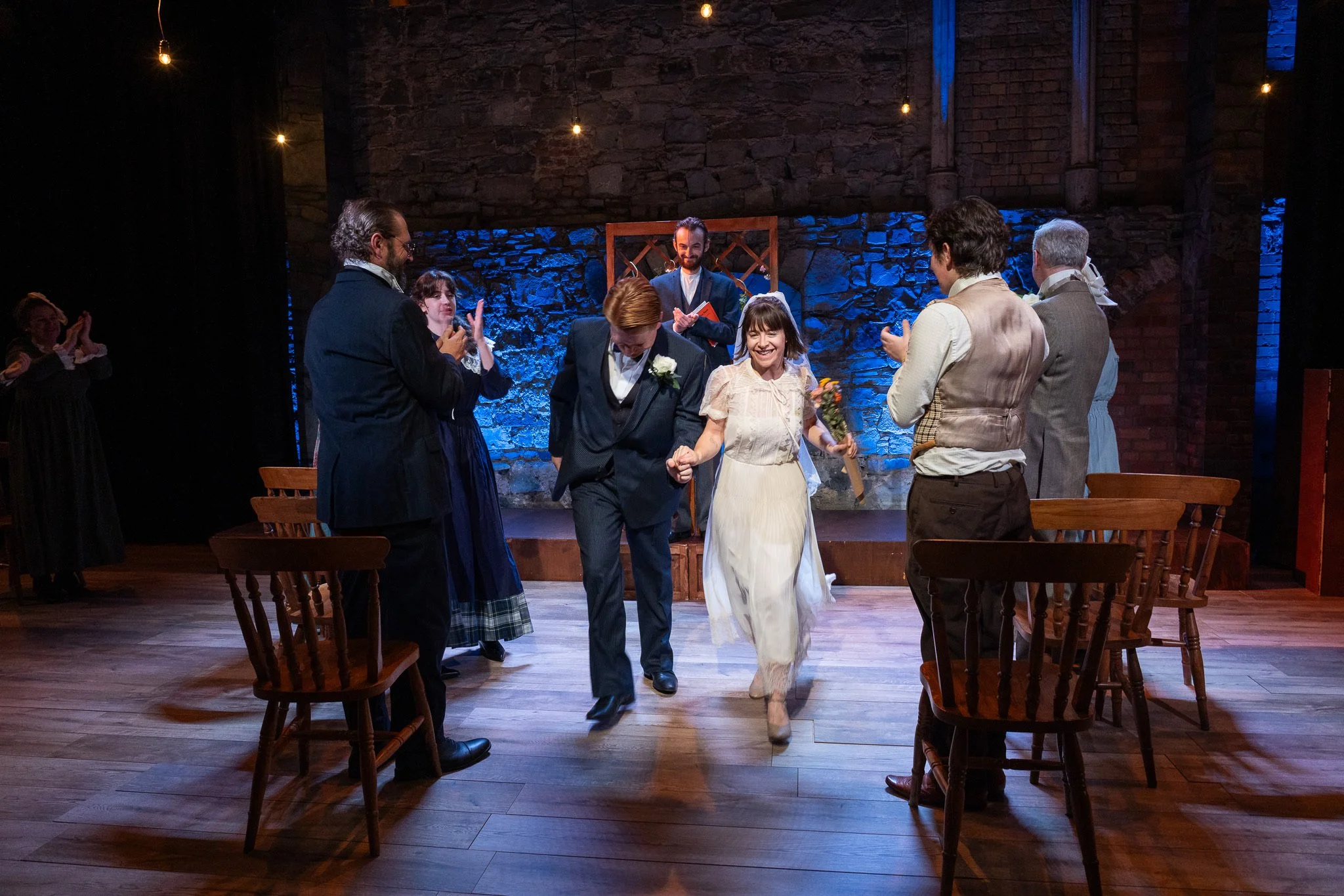 A group of people dressed in vintage clothing are gathered on a stage with a stone wall background, some clapping and one person holding flowers, during a theatrical performance or ceremony.