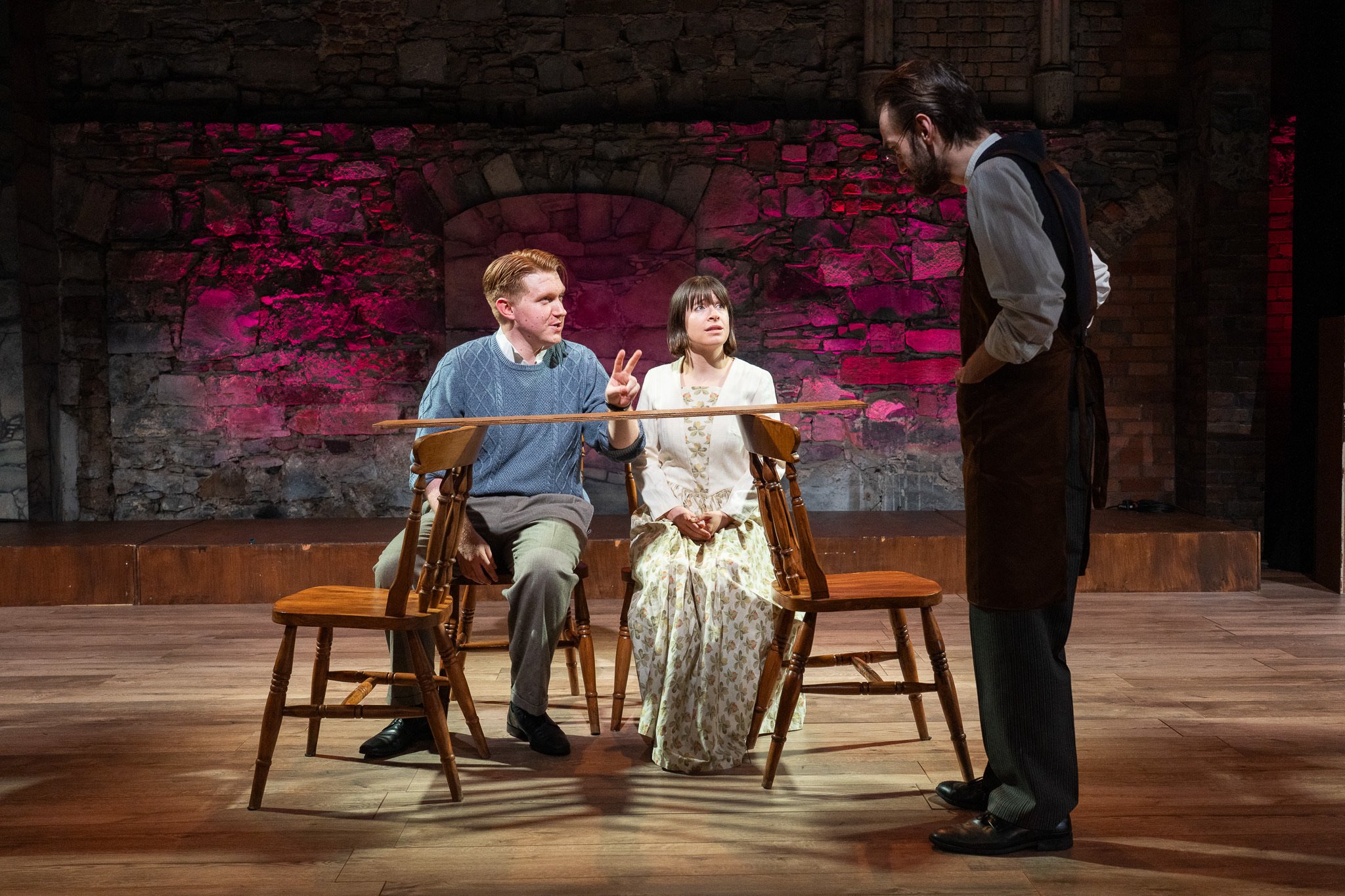 Three actors in costume performing a scene on stage with a stone wall background, one man standing and two seated, one making a peace sign, with a person standing in front of them.