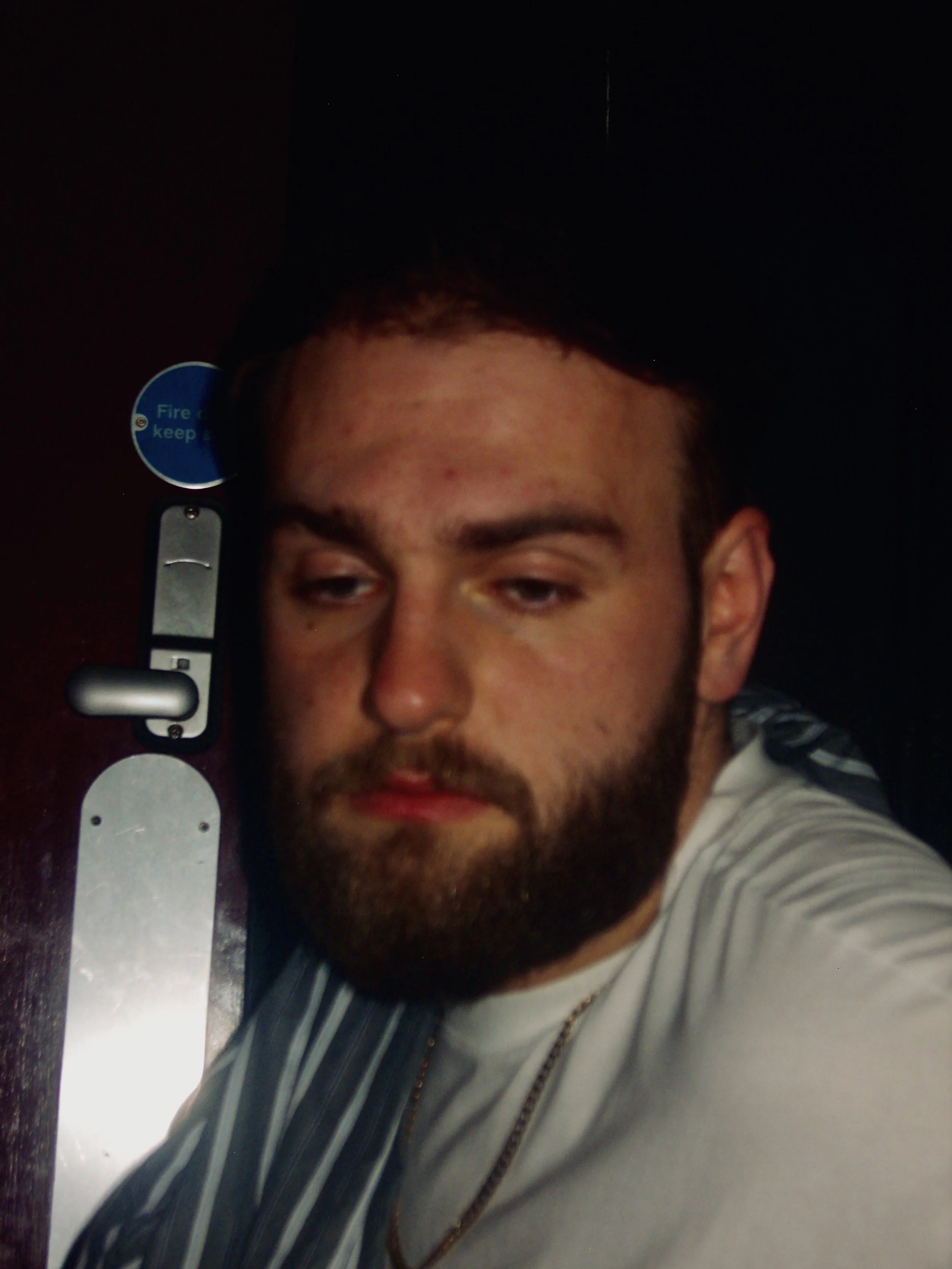 A young man with short dark hair and a beard taking a selfie, wearing a striped jacket and a white shirt, lying on a dark-colored surface.