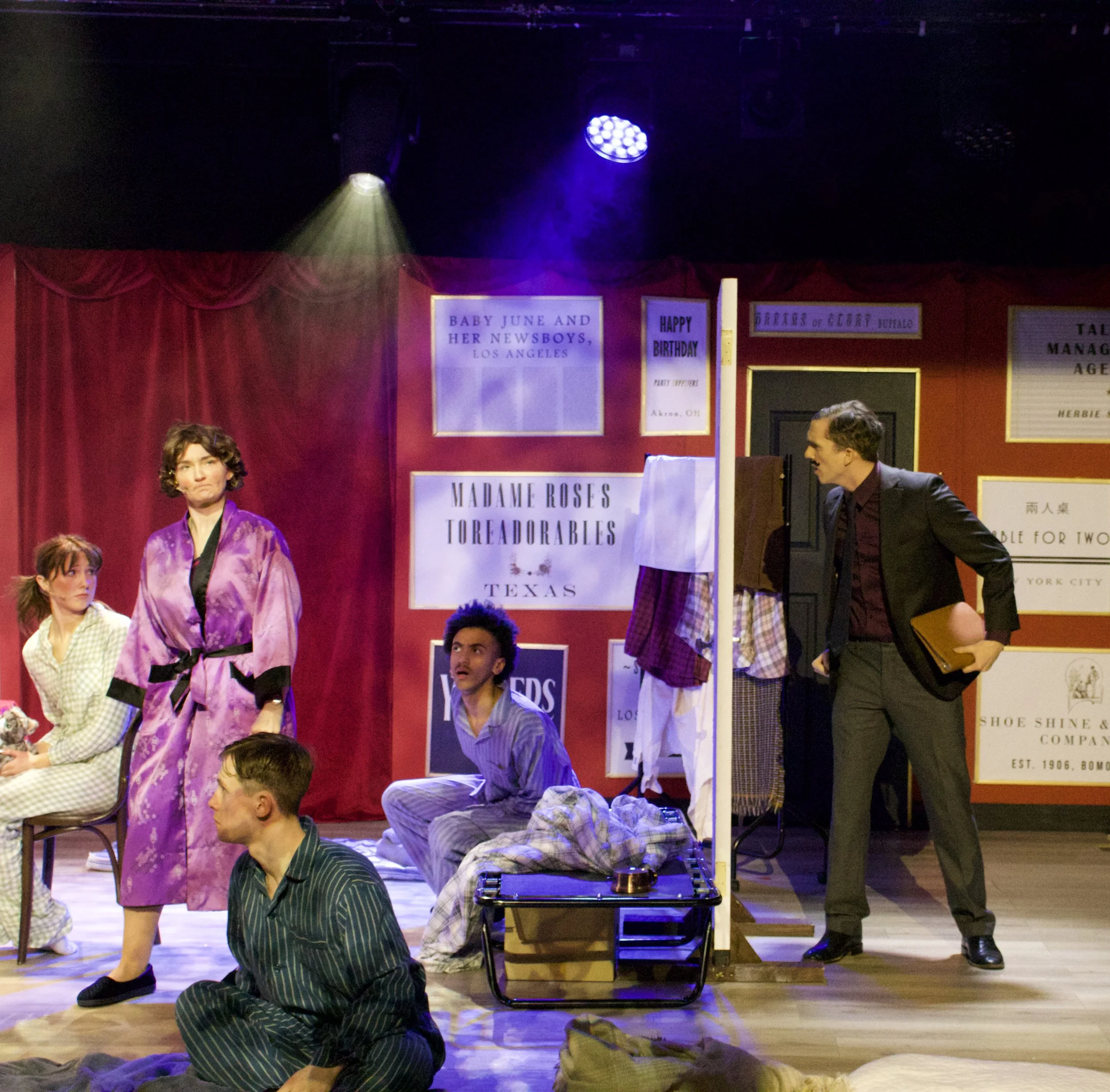 The stage of a theater with purple curtains and decorative signs, where actors are performing a play. Several actors are in costume, with some standing and others seated on the stage. Stage lighting includes purple and yellow spotlights.