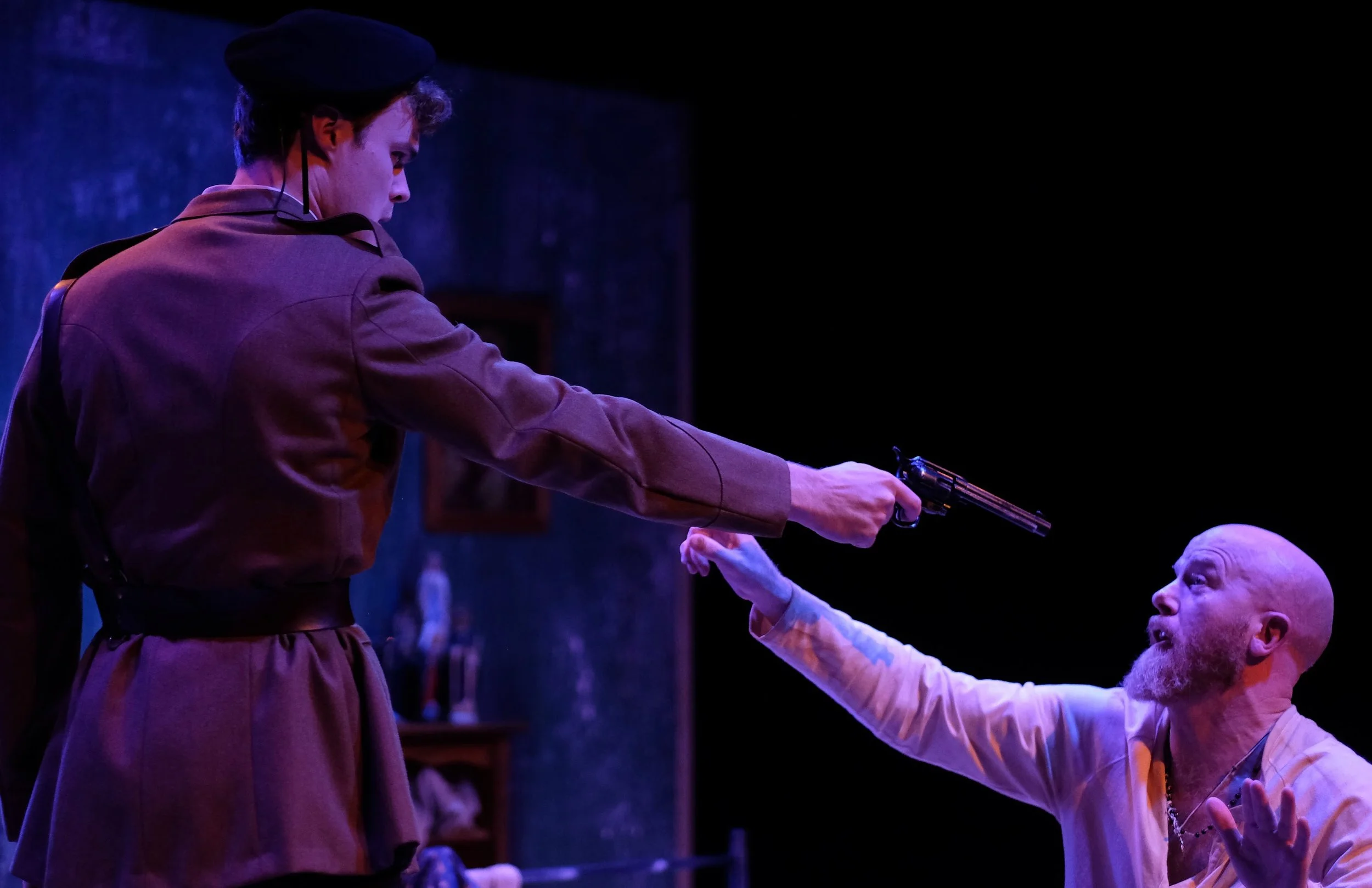 A man in a police uniform aiming a gun at a young woman, who is reaching out to him with her hand in a dimly lit room.
