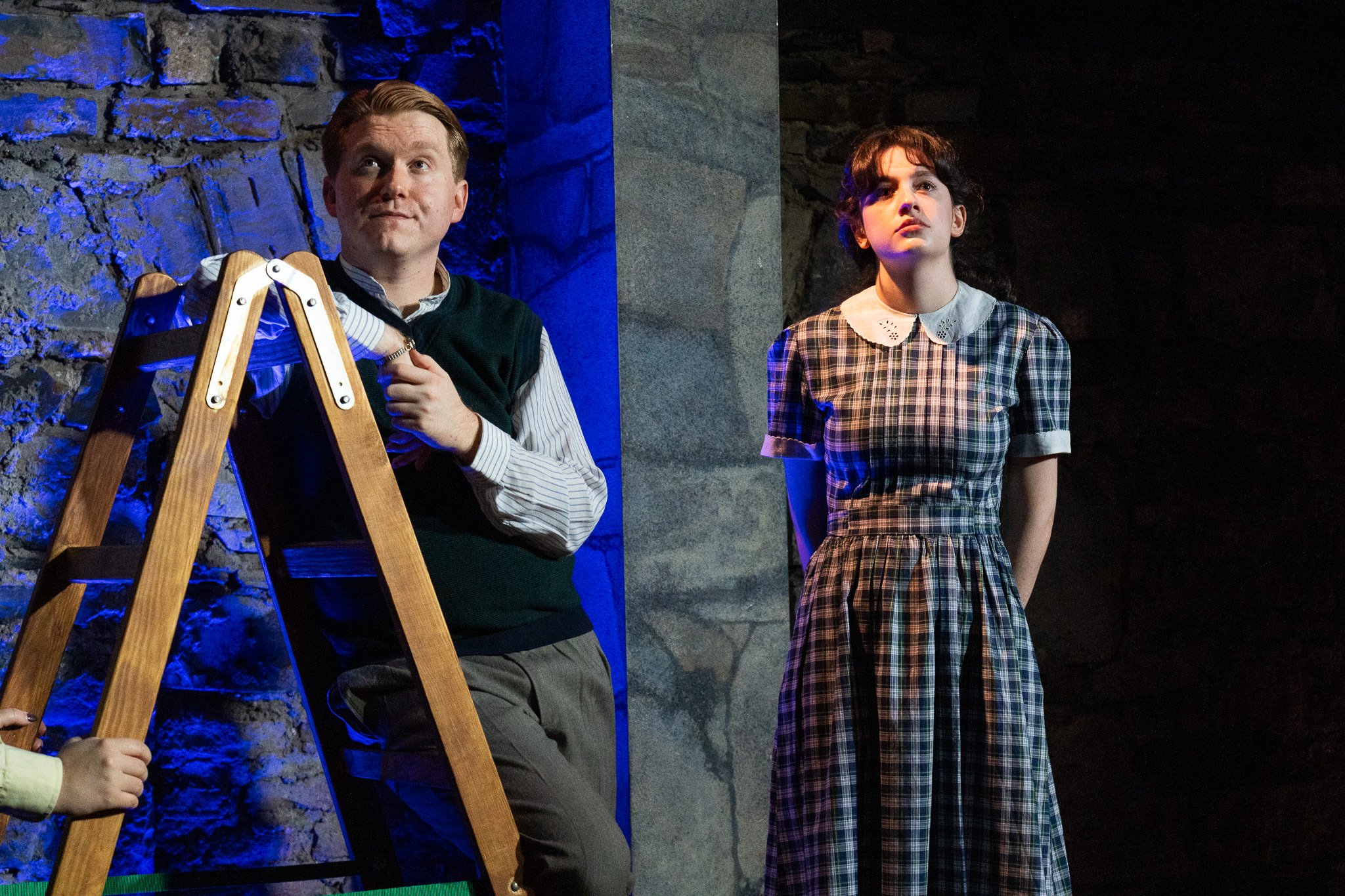 A scene from a theatrical performance with two actors on stage. The male actor on the left has light skin, red hair, and is sitting on a wooden ladder, wearing a dark vest over a striped shirt. He is holding a hand to his chest and looking upward. Th