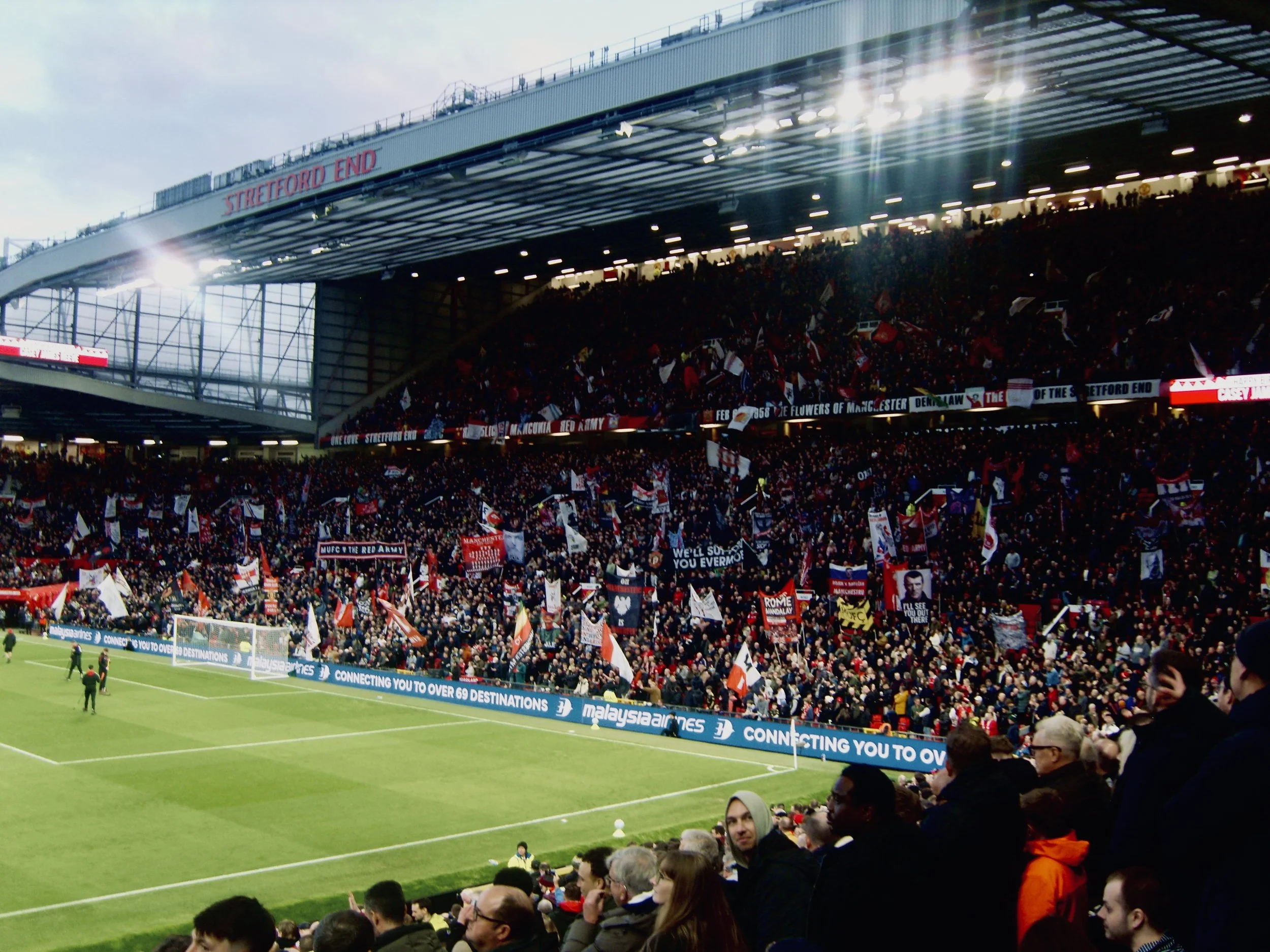 Crowd inside a soccer stadium with banners, flags, and fans watching a match on the field, with a large overhang displaying 'Stretford End'.