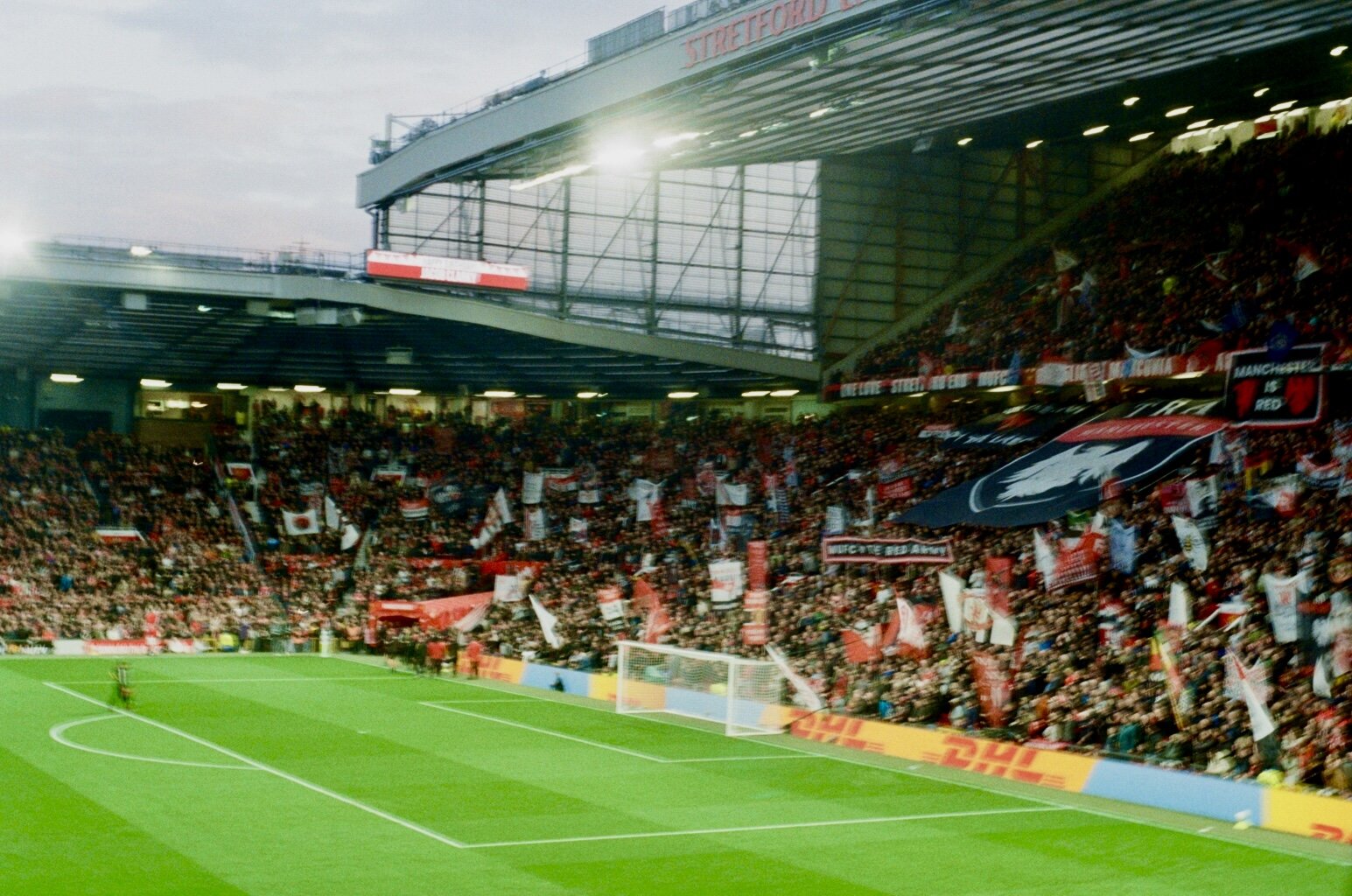 A packed soccer stadium stands filled with fans waving flags and banners, some with a red and white theme, under a partially cloudy sky.