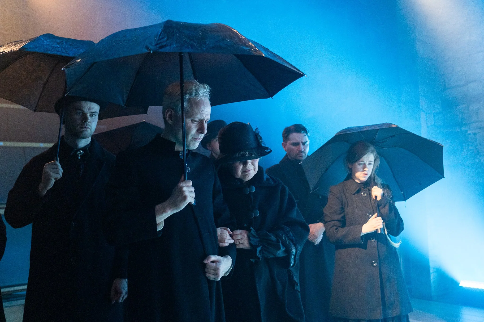 A group of people standing closely together holding black umbrellas, some with their heads bowed, appears to be in a somber or memorial gathering.
