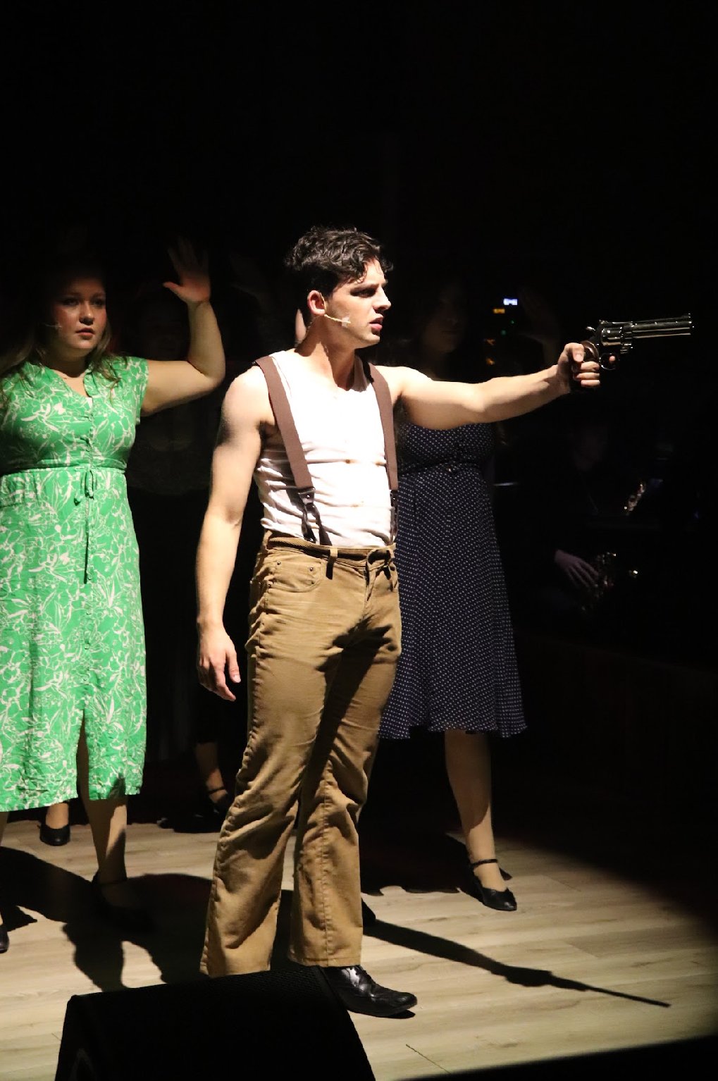 Stage performers in costumes, with a male actor holding a revolver, during a theatrical play.
