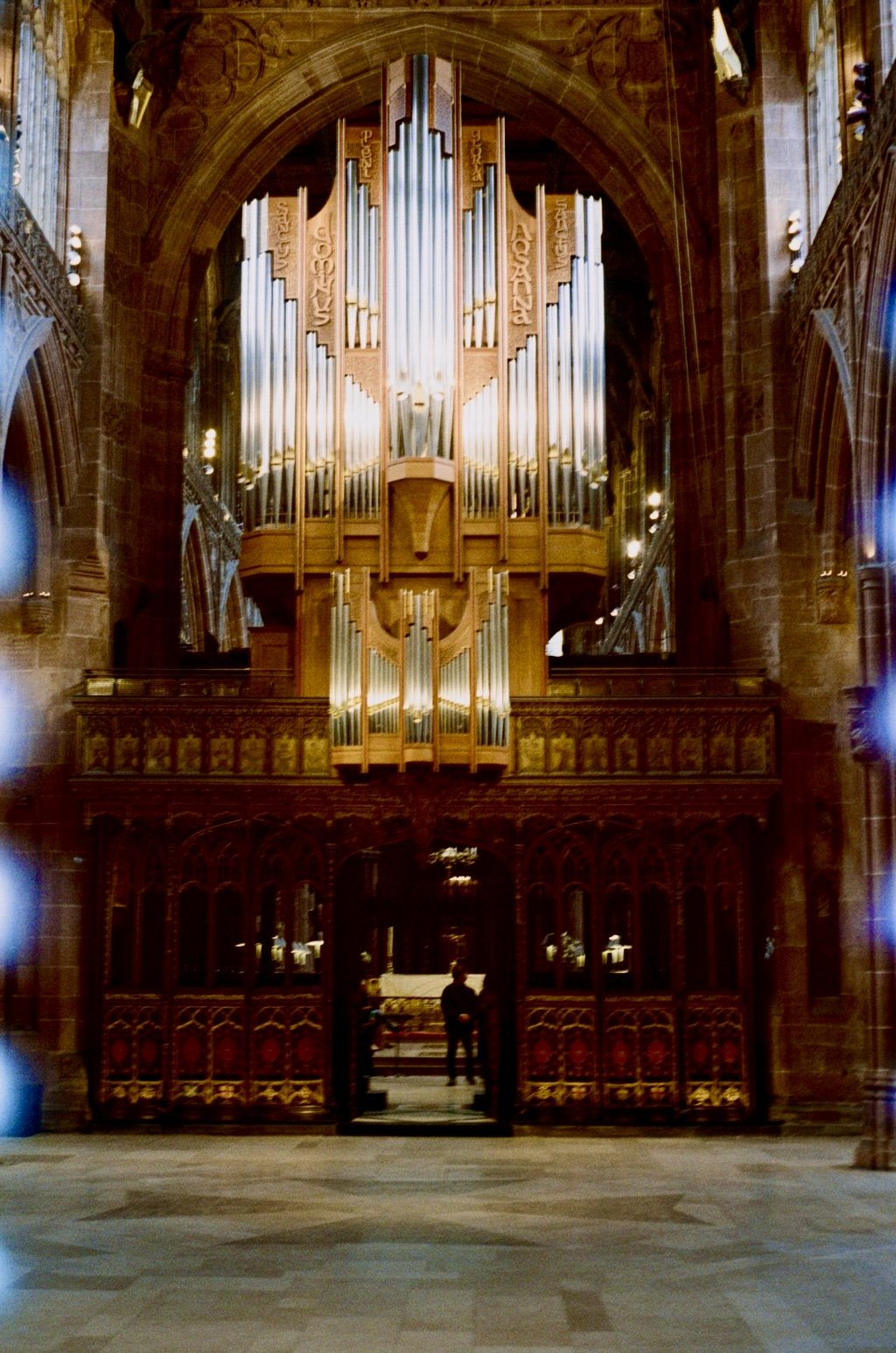 A large pipe organ with numerous metal pipes in a church or cathedral, decorated wooden carvings, and a person standing near the organ.