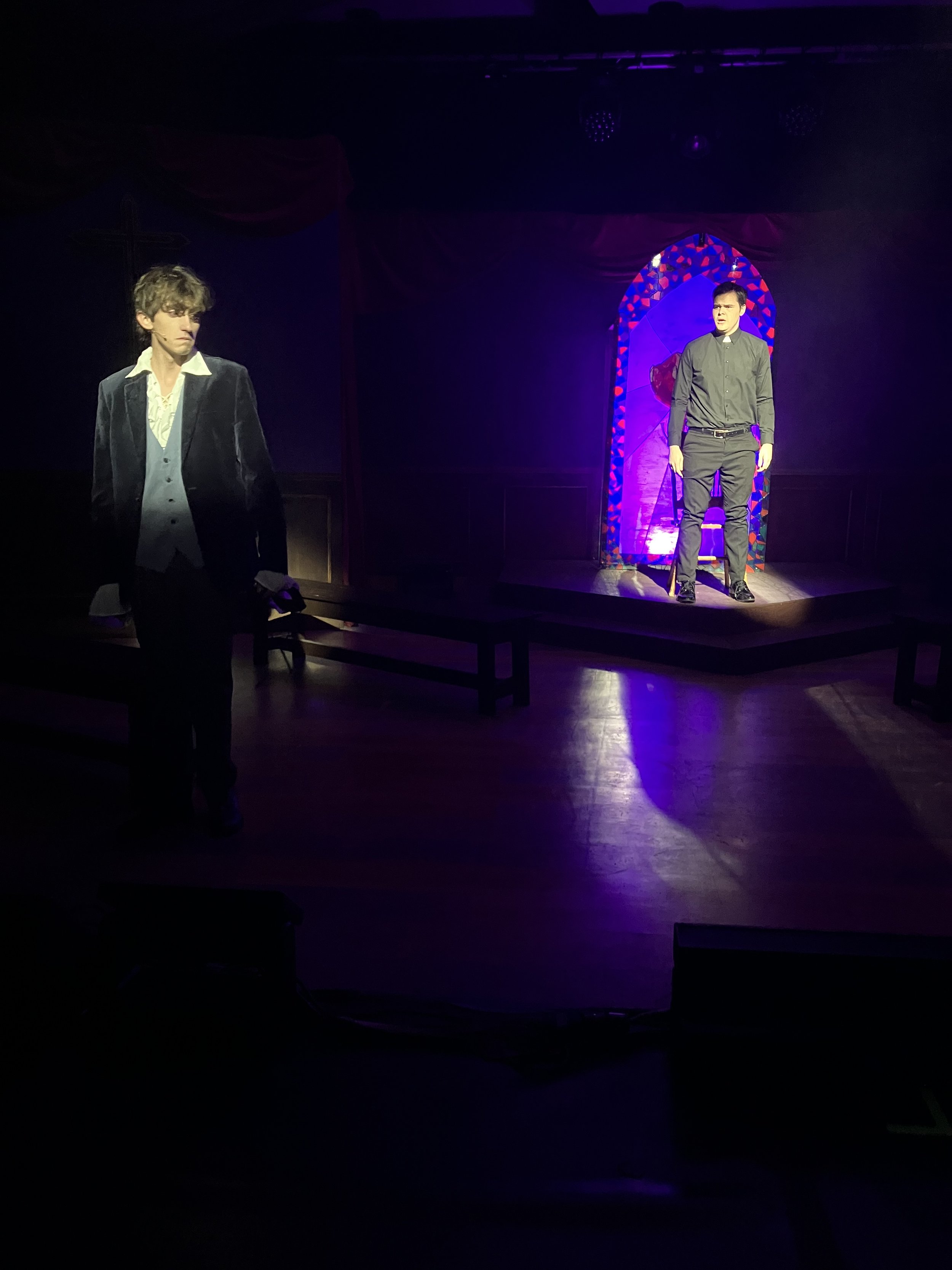 Two men on a stage with a colorful stained-glass-style backdrop, one dressed as a priest and the other in a suit, under purple stage lighting.