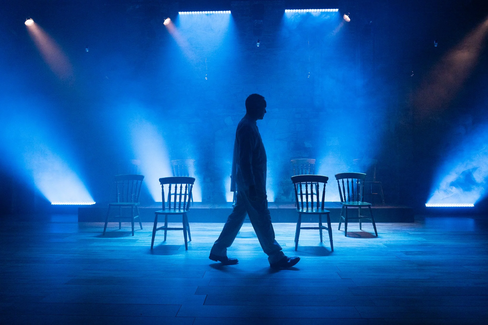 A person walking across a stage with a blue spotlight, four chairs lined up behind them, a brick wall in the background, and stage lighting above.