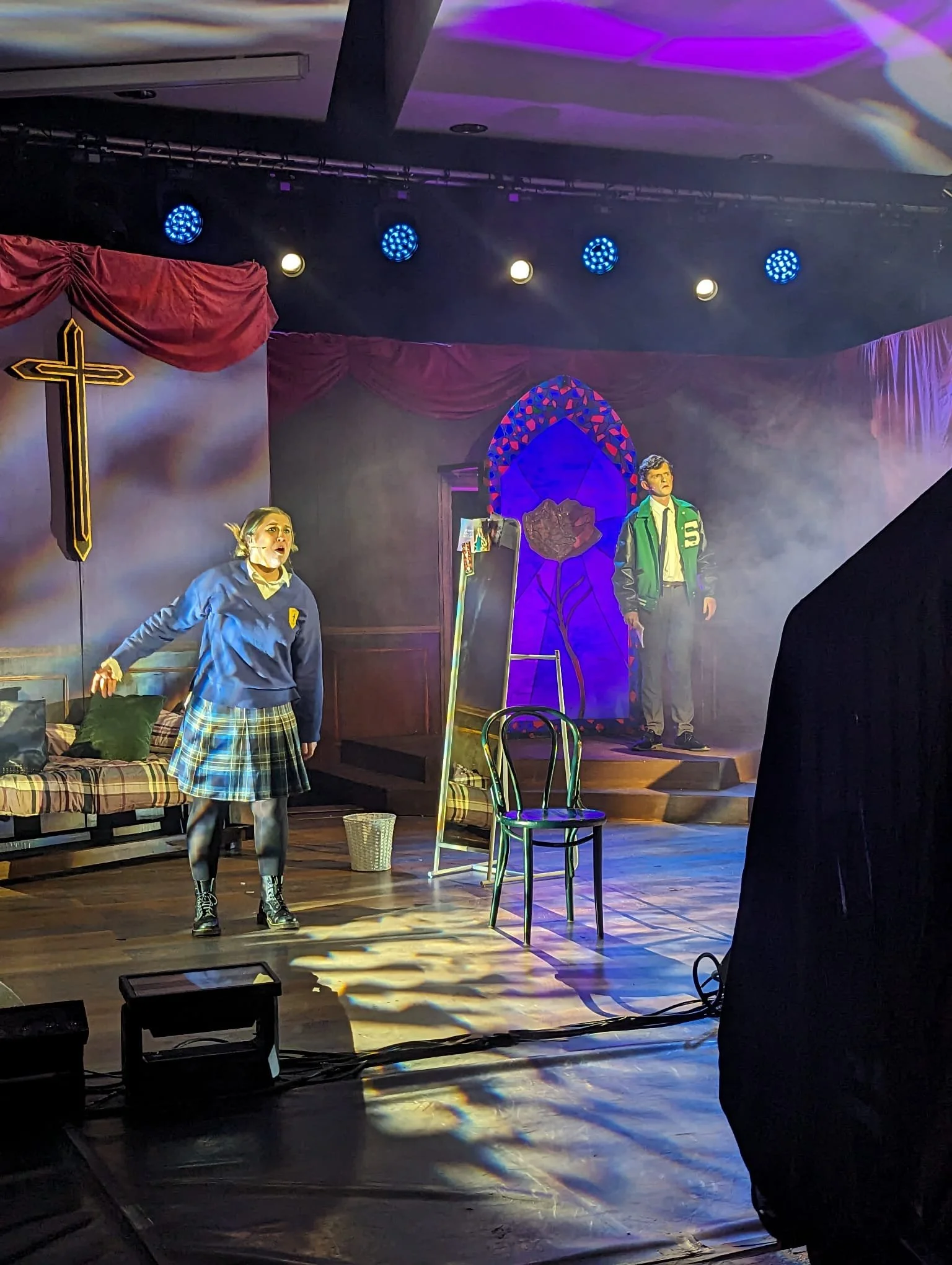 Stage with two actors performing. Female actor in a blue jacket and plaid skirt on the left, male actor in a green letterman jacket on the right. Stage is decorated with a cross, red curtains, a stained glass window, and a chair. Theater lighting cre
