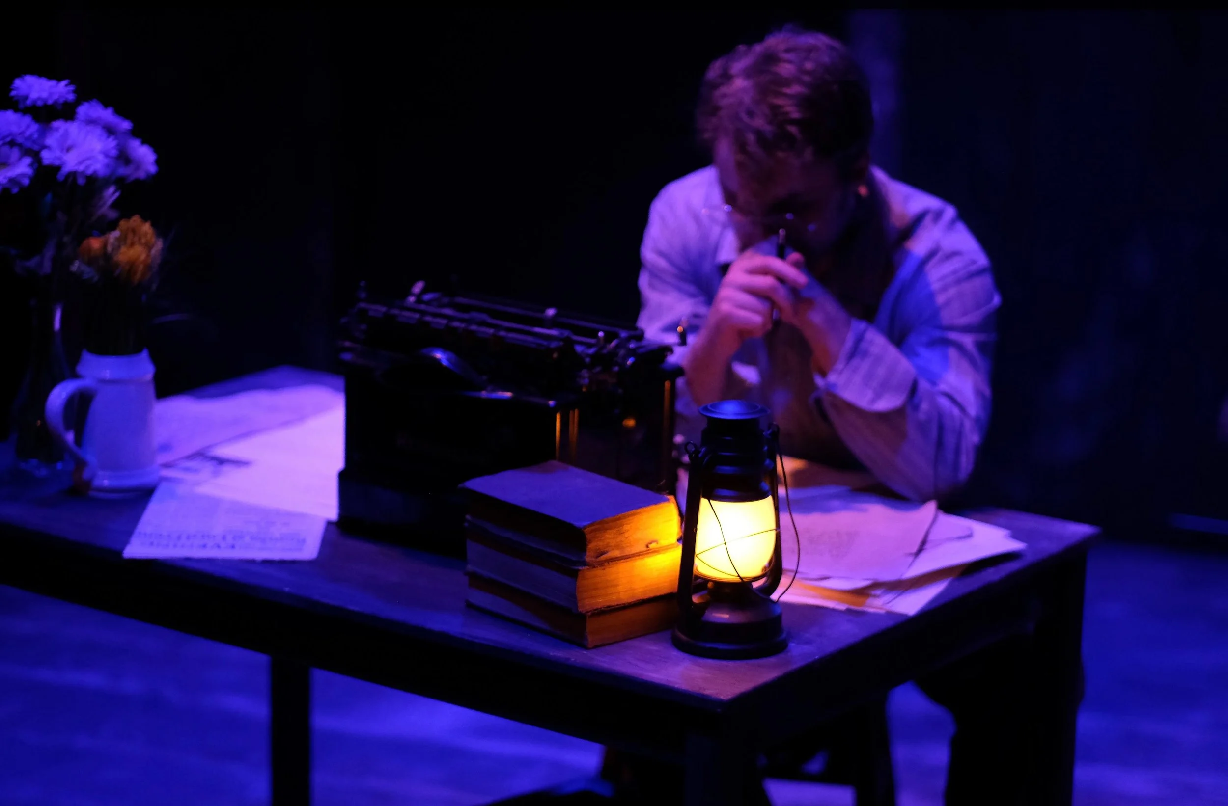A person sitting at a table in a dark room illuminated by a small lantern, with flowers and papers on the table.