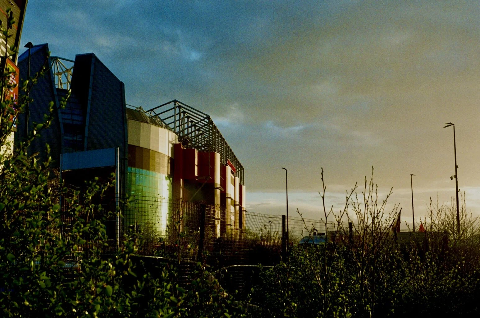 A modern building with reflective glass windows and a colorful exterior in the sunlight, surrounded by bushes and a fence, under a partly cloudy sky.