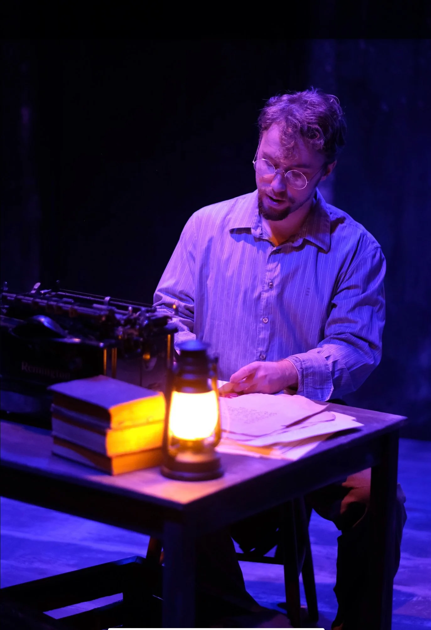 A man with glasses and curly hair wearing a striped shirt, sitting at a table with books, a notebook, and papers, illuminated by a warm lamp on the table, with a dark background and purple lighting.