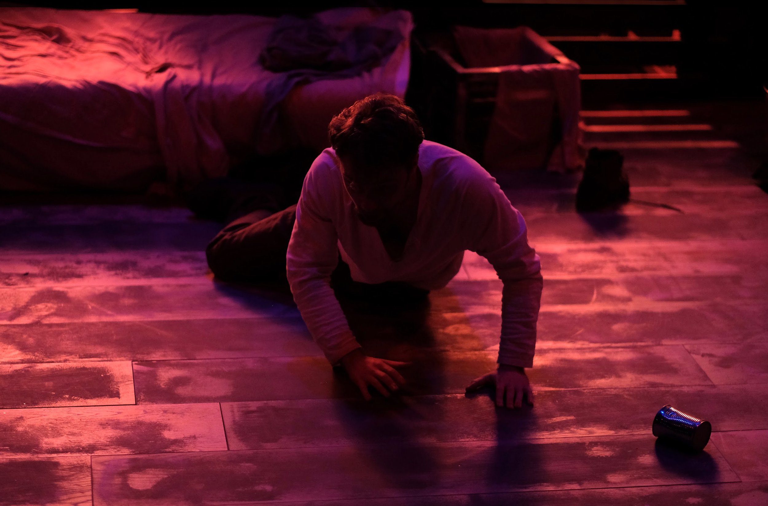 Person on hands and knees on a wooden floor under colorful stage lighting, with a bed, clothes, and furniture in the background.