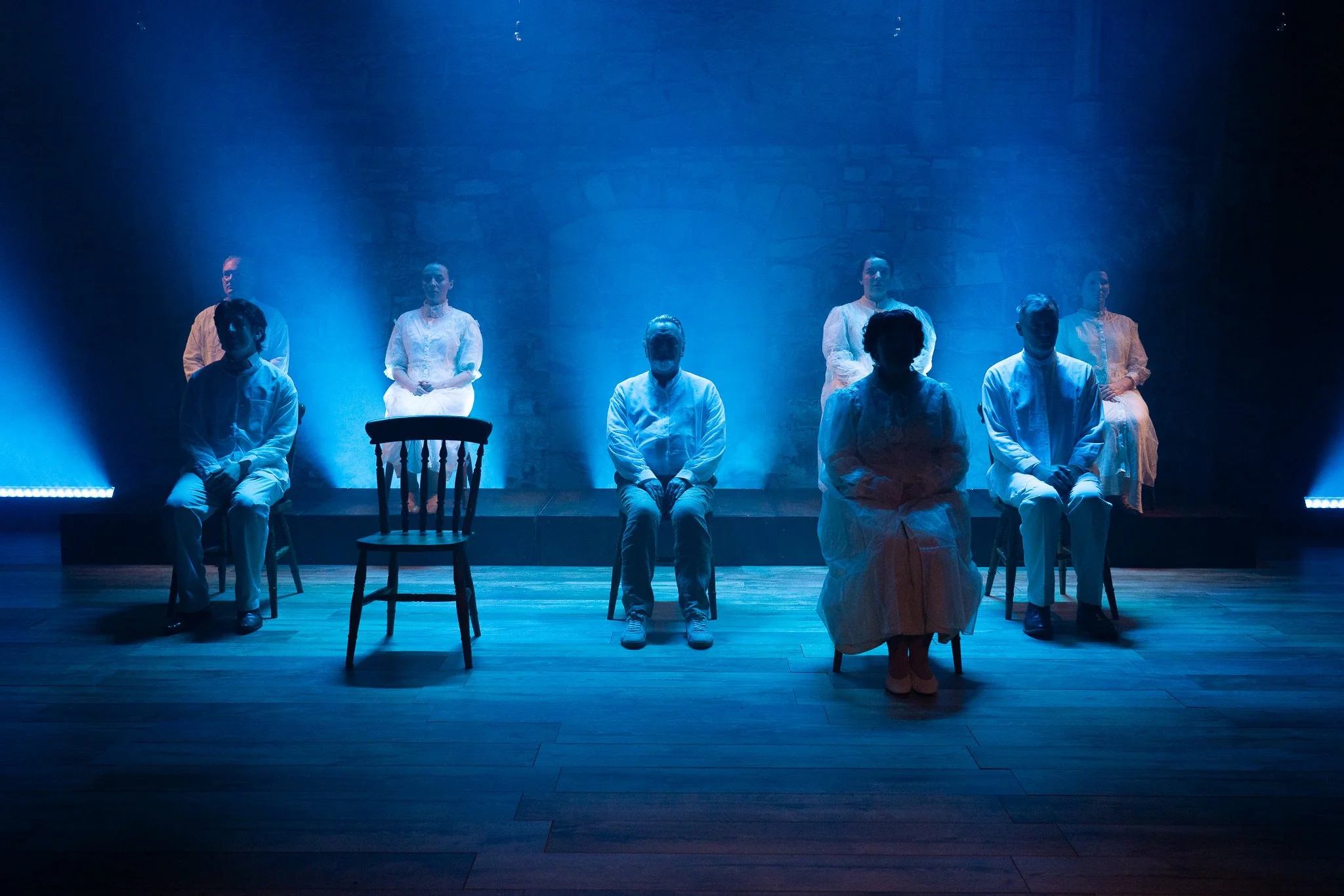 Severals people dressed in white sitting on chairs on stage with blue lighting and a brick wall in the background.