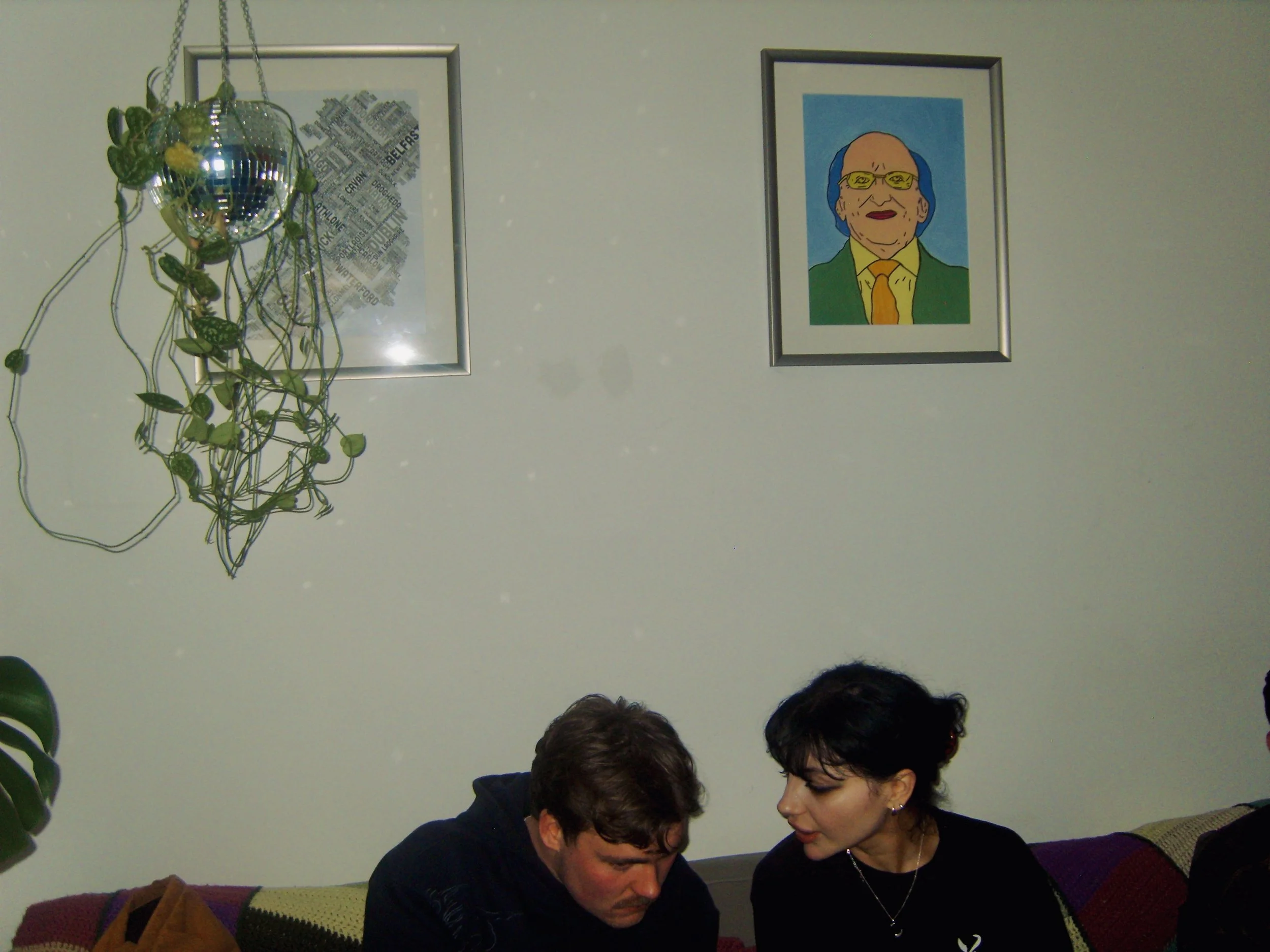 Two people sitting closely on a sofa, engaged in conversation. There are framed pictures on the wall behind them, including a cartoon-like portrait of an elderly person with glasses and a colorful background, and a decorative hanging object resemblin