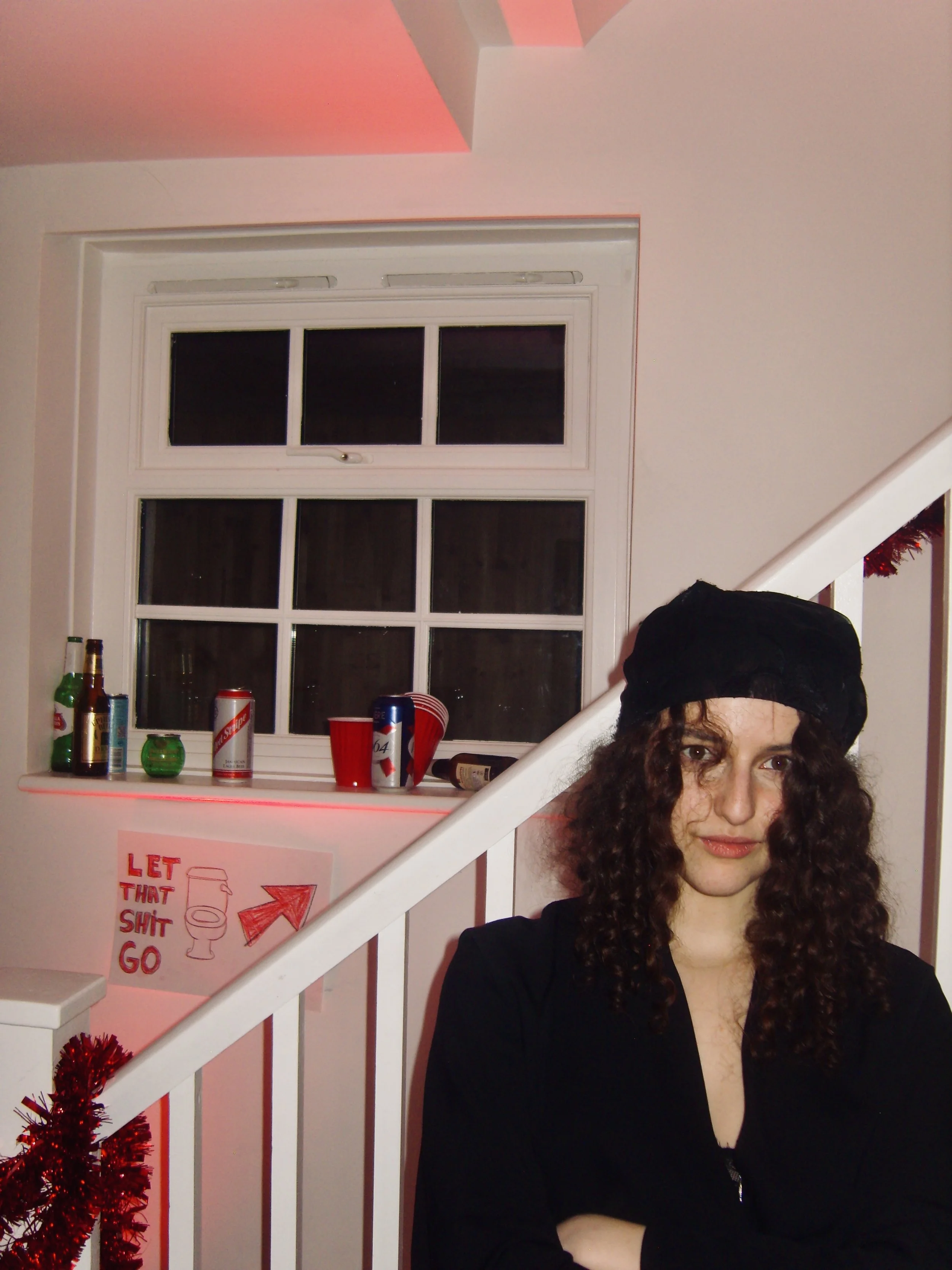 A woman with curly hair wearing a black shirt and black head covering sitting on a staircase inside a house with a pink wall. There is a window with white frame above the staircase. Bottles and red cups are on the windowsill. A red mini Christmas tre