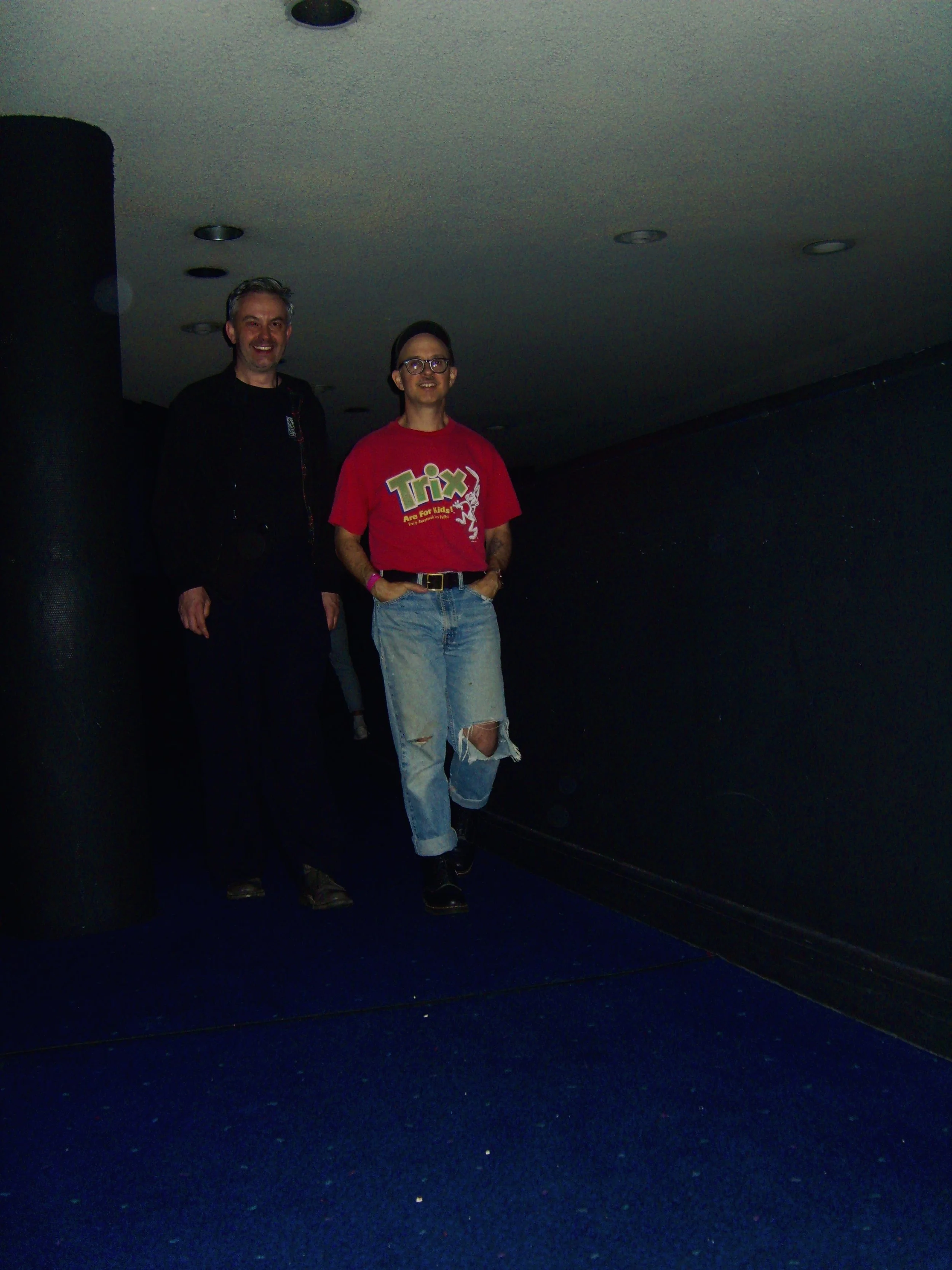 Two men standing against a dark wall, both smiling. The man on the left wears a black jacket, and the man on the right wears a red T-shirt with a logo that says "Trix" and ripped jeans.