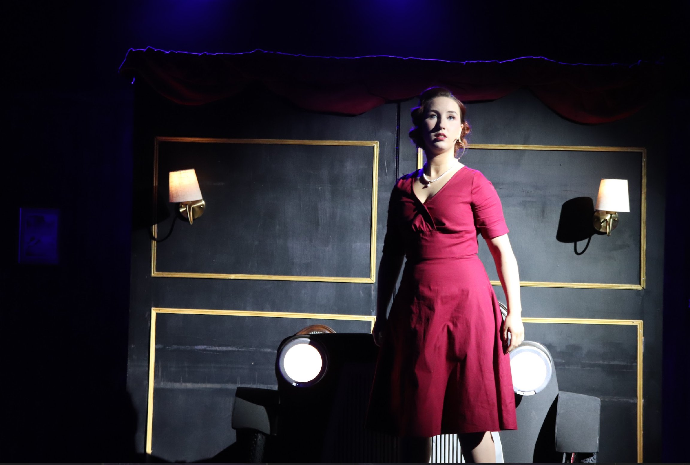 A woman in a vintage red dress stands on a stage with dark walls, framed gold borders, and two wall-mounted lamps with shades, illuminated by stage lighting.