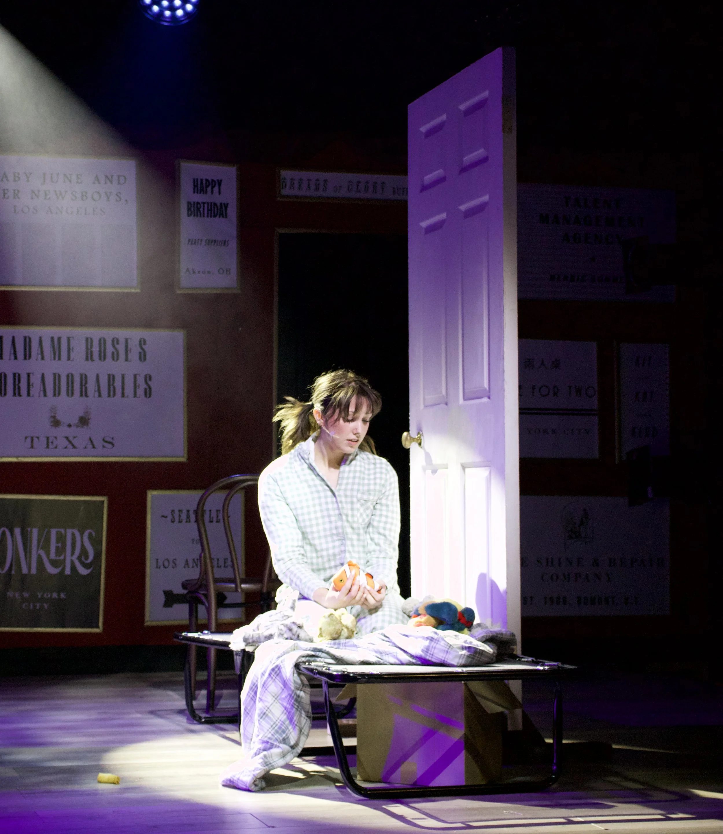 A woman in plaid pajamas sitting on a hospital gurney surrounded by stuffed animals, holding a stuffed animal, with posters on the wall behind her and a partially open door lit with purple light.
