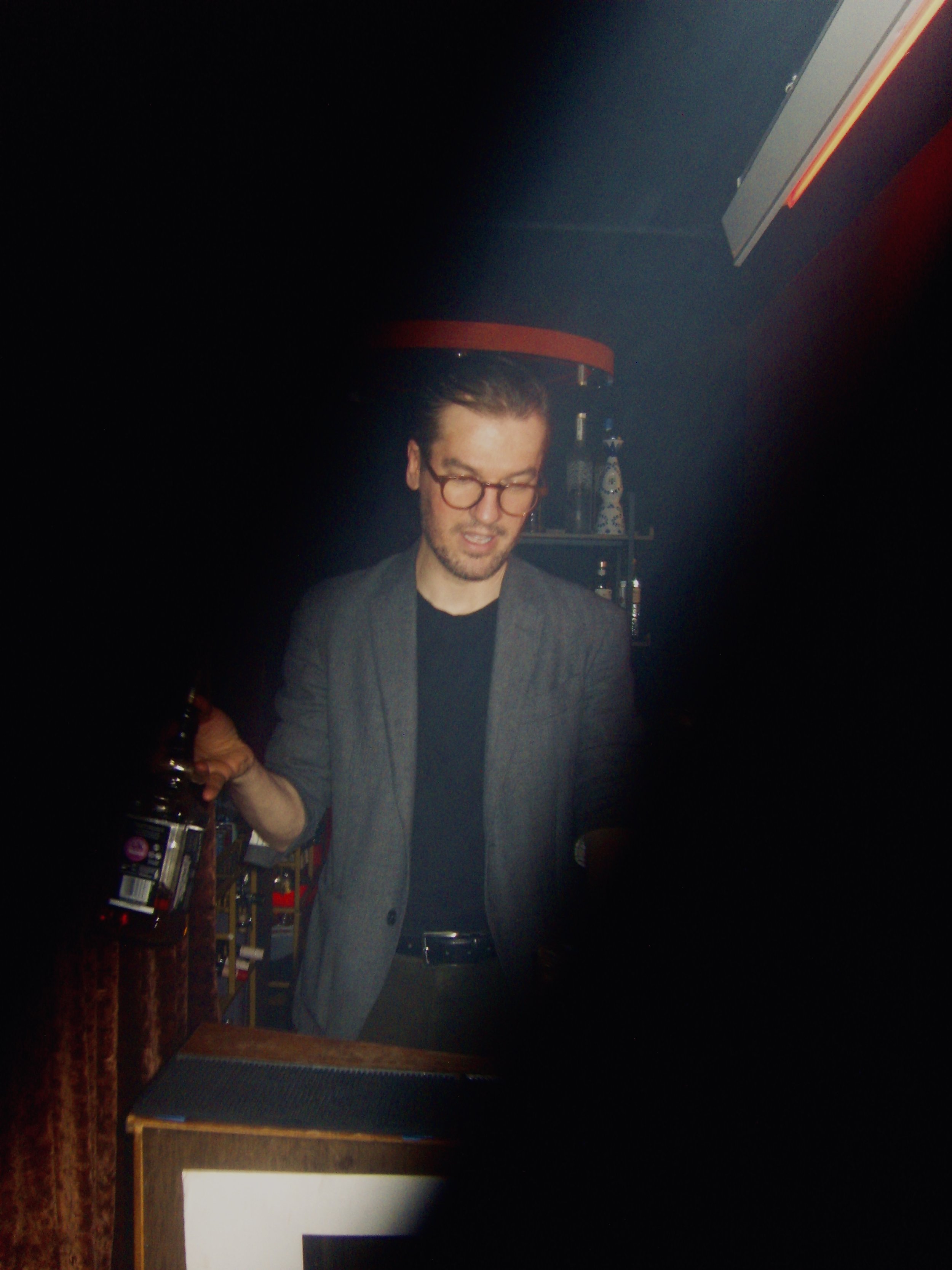 A man with glasses and a beard wearing a gray jacket and black shirt, holding a bottle, standing in a dimly lit room with shelves of bottles in the background.