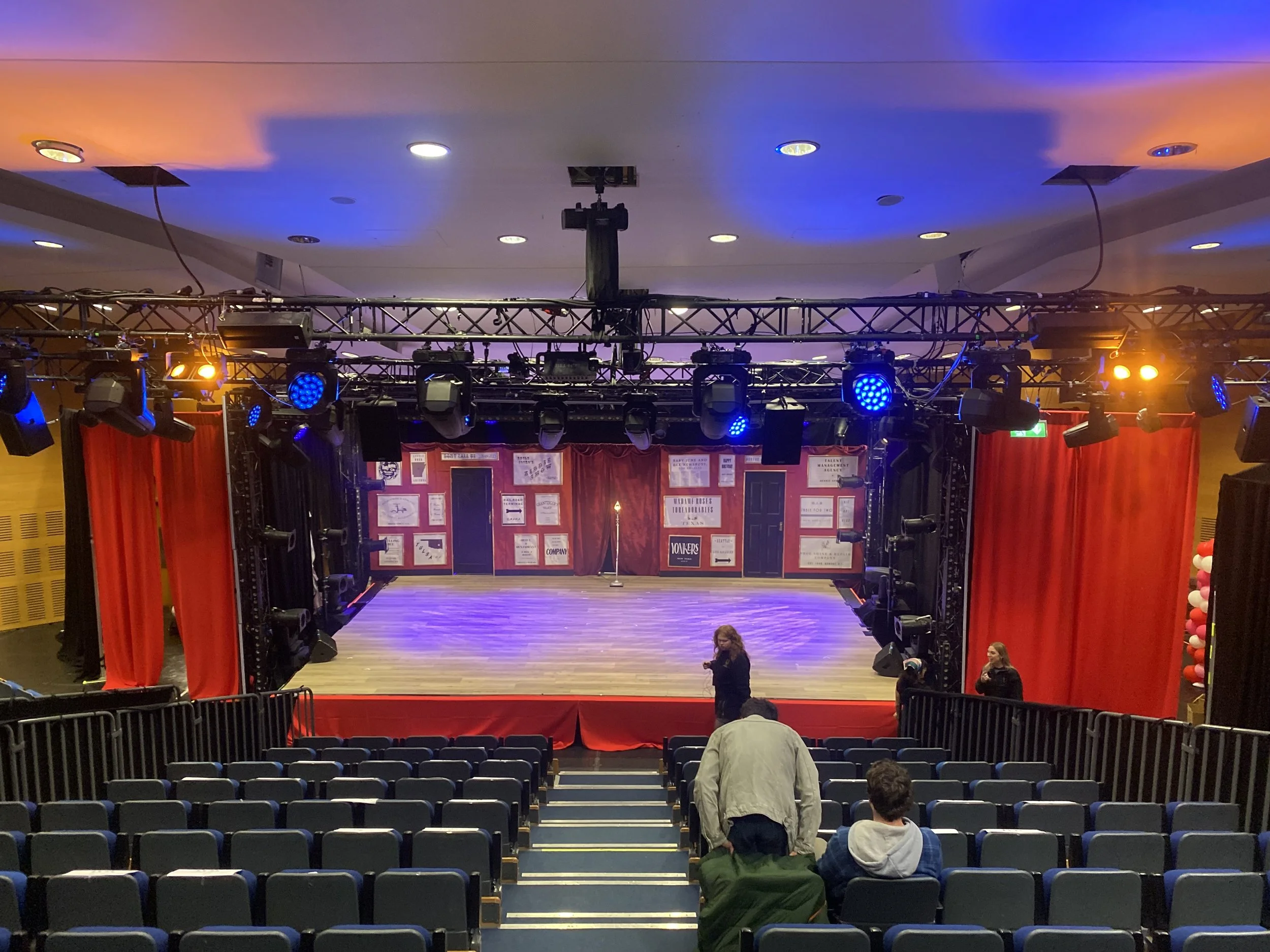 An empty stage set up with numerous stage lights, red curtains, and a backdrop with posters or signs. Seating in front with a few people preparing or waiting.