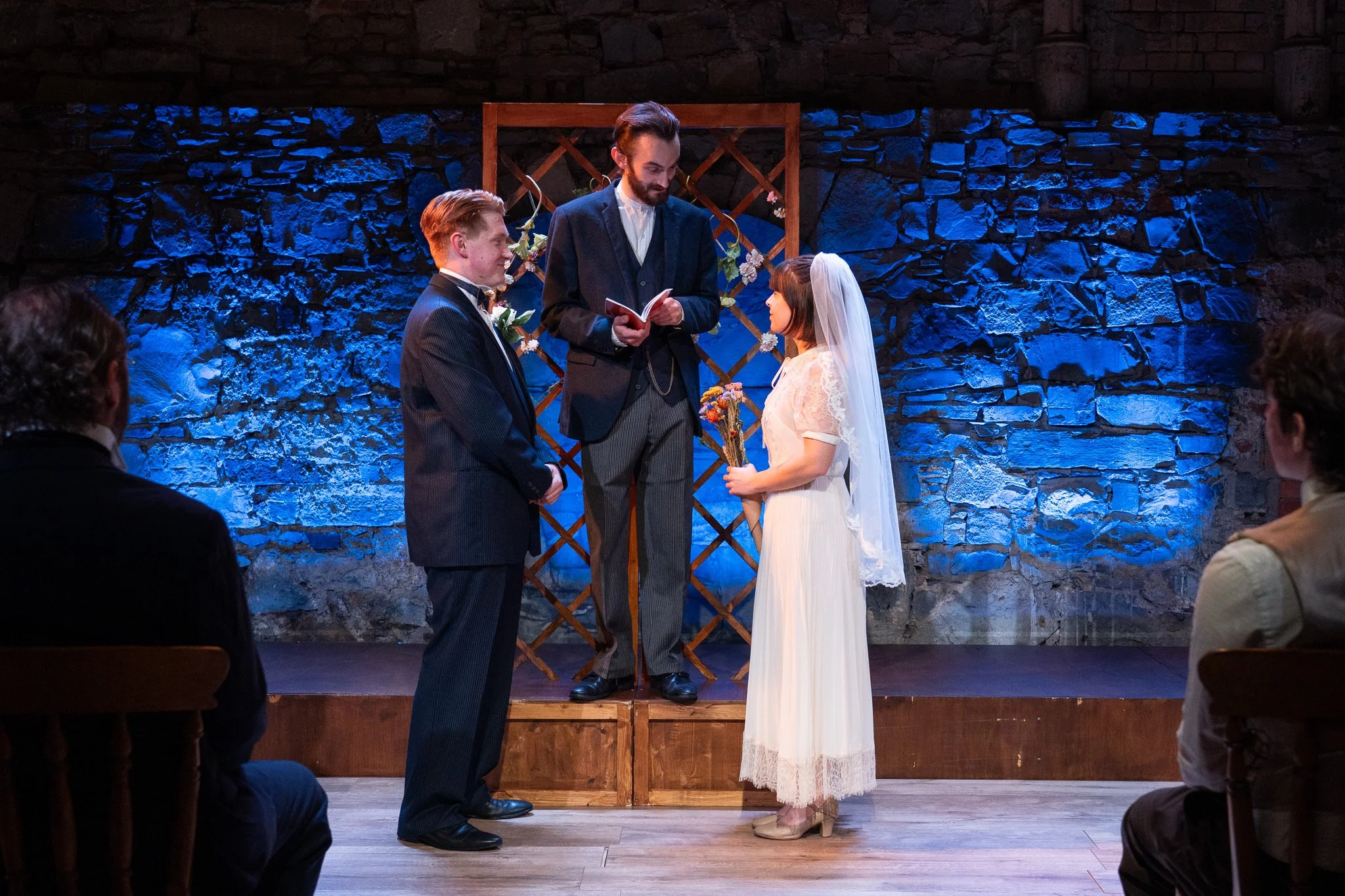 A wedding ceremony with a couple exchanging vows on a stage with a stone wall backdrop, officiant reading from a book, guests seated facing the couple.