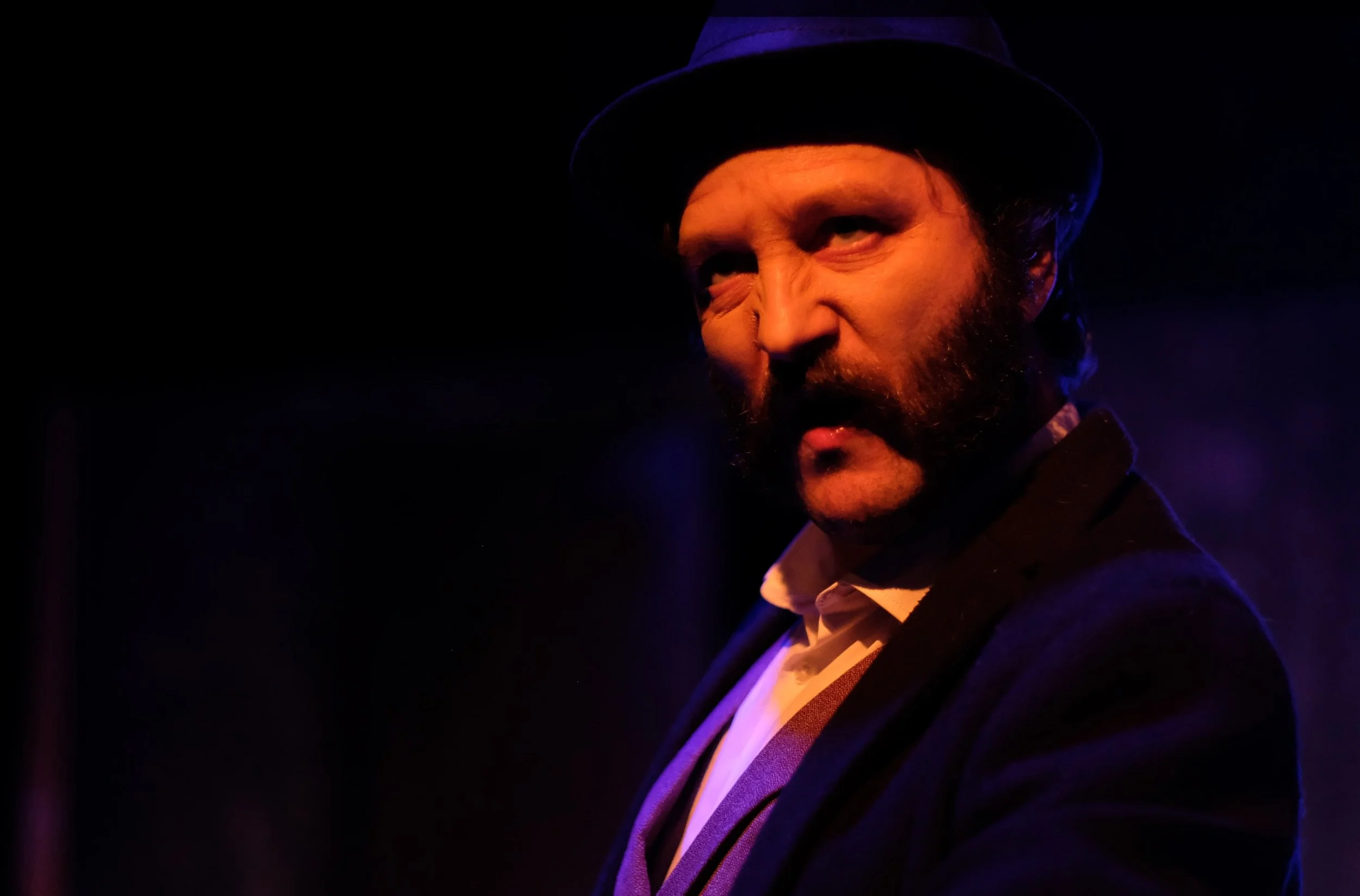 A man with a beard wearing a dark hat and suit, illuminated by dramatic purple and orange lighting.