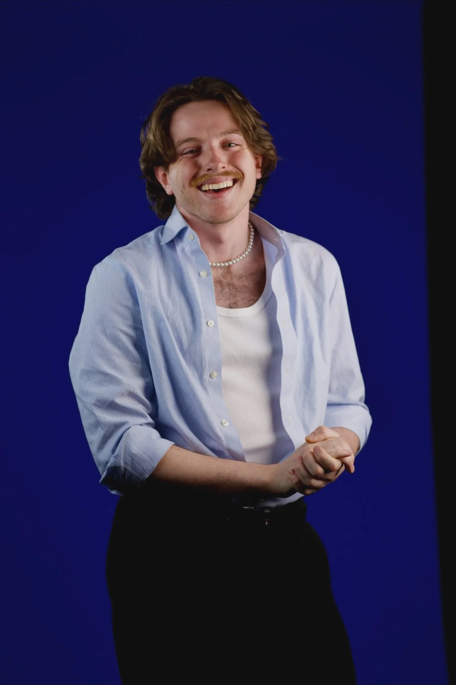 A young man with brown wavy hair, a mustache, and a beard, smiling, wearing a white t-shirt, a light blue striped shirt, a pearl necklace, and black pants, standing against a blue background.