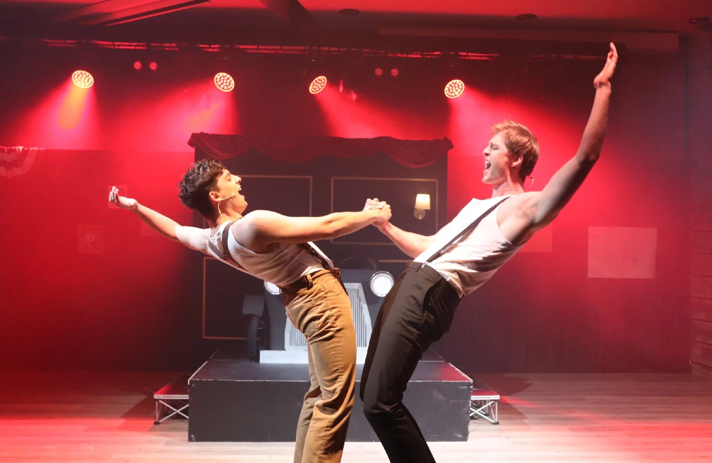 Two performers on stage, holding hands, dancing passionately in front of red stage lights with a dark background and a red curtain overhead.